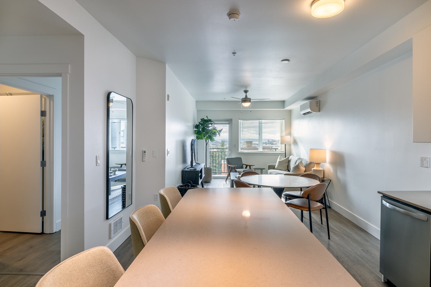 Bright kitchen and dining area in an apartment with modern white cabinets, a tiled backsplash, wood floors, and a cozy bar-height table with stools.