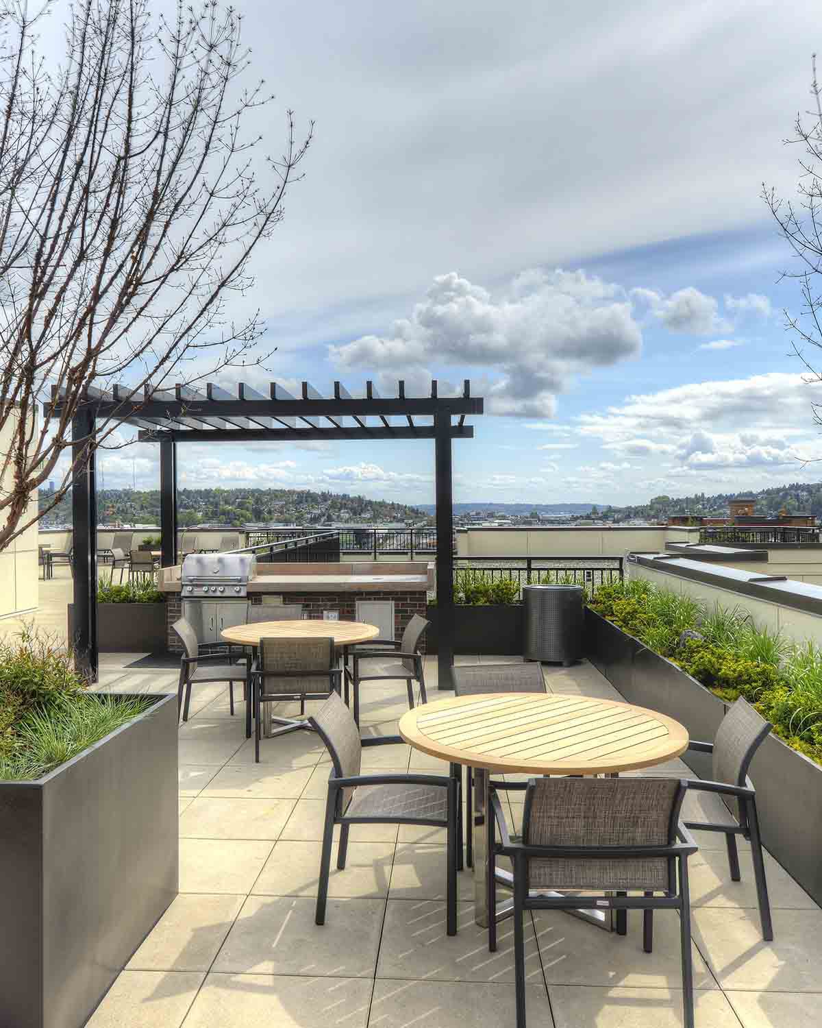 Rooftop apartment building terrace featuring outdoor dining tables, a pergola with a BBQ grill, lush planters, and scenic neighborhood views.