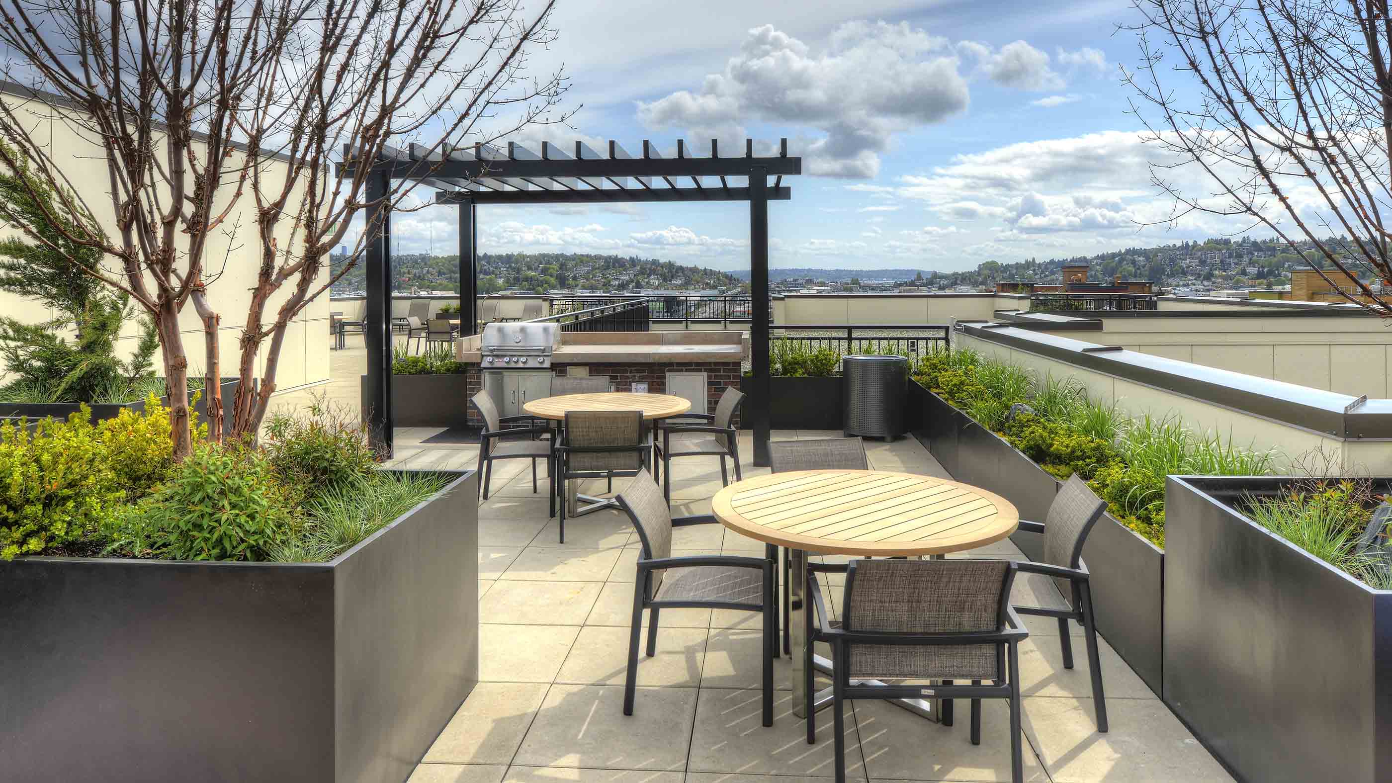 Rooftop apartment building terrace featuring outdoor dining tables, a pergola with a BBQ grill, lush planters, and scenic neighborhood views.