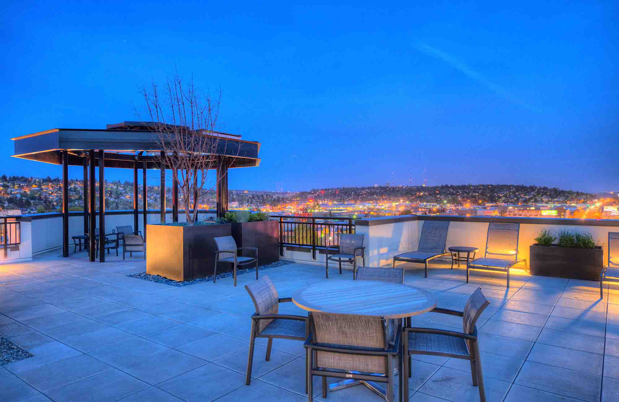 Rooftop terrace at dusk with modern seating, pergola, and planter boxes, overlooking a glowing Ballard cityscape under a deep blue sky.