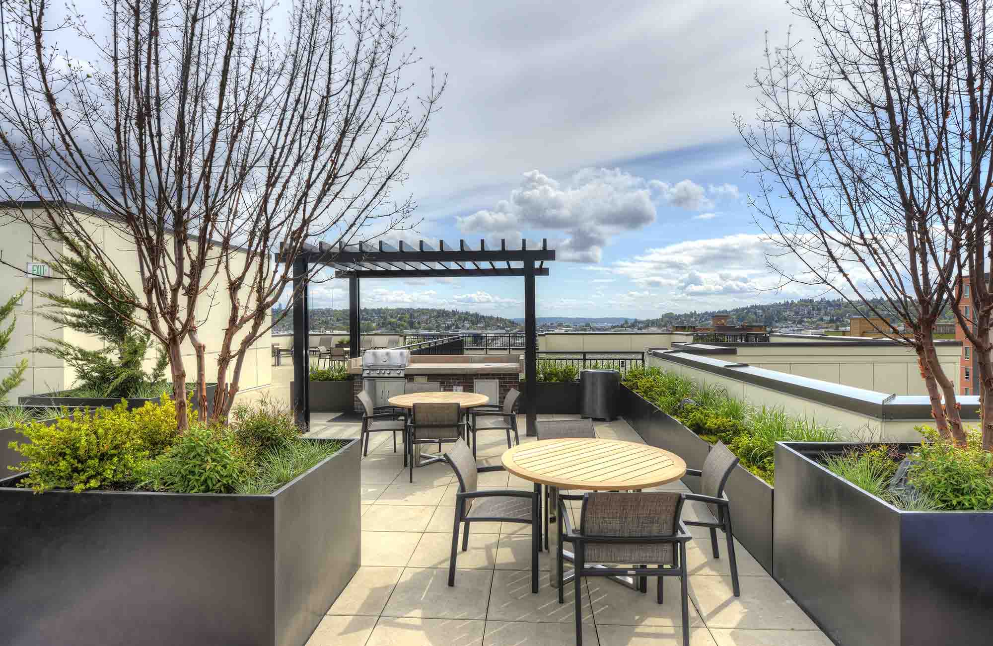 Rooftop terrace with modern outdoor seating, barbecue grill, and pergola, surrounded by raised planters and offering distant views of hills under a partly cloudy sky.