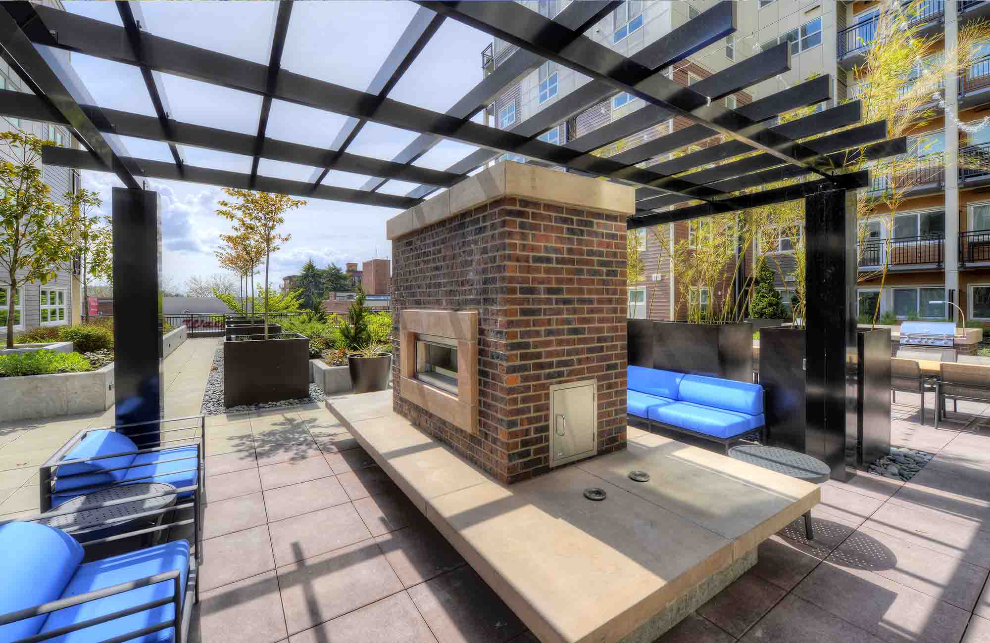 Daytime view of The Waymark Apartments’ courtyard with a sleek, brick-built, double-sided outdoor fireplace under a pergola, featuring blue cushioned lounge seating, modern planters, and nearby grilling and dining areas.