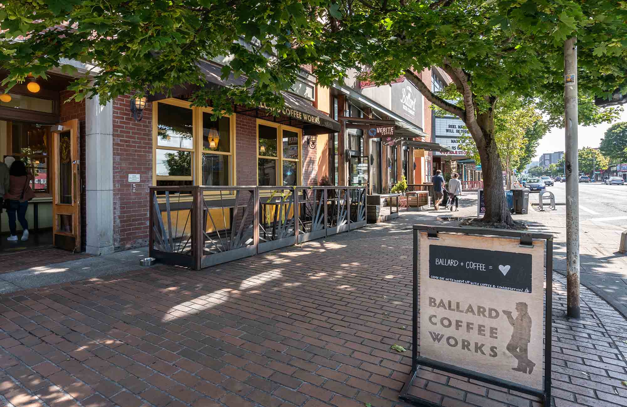 Street-level view of Ballard Coffee Works, a cozy brick café with yellow-trimmed windows and a wooden patio, located on a tree-lined sidewalk with a sandwich board sign in the foreground that reads “Ballard Coffee Works.”