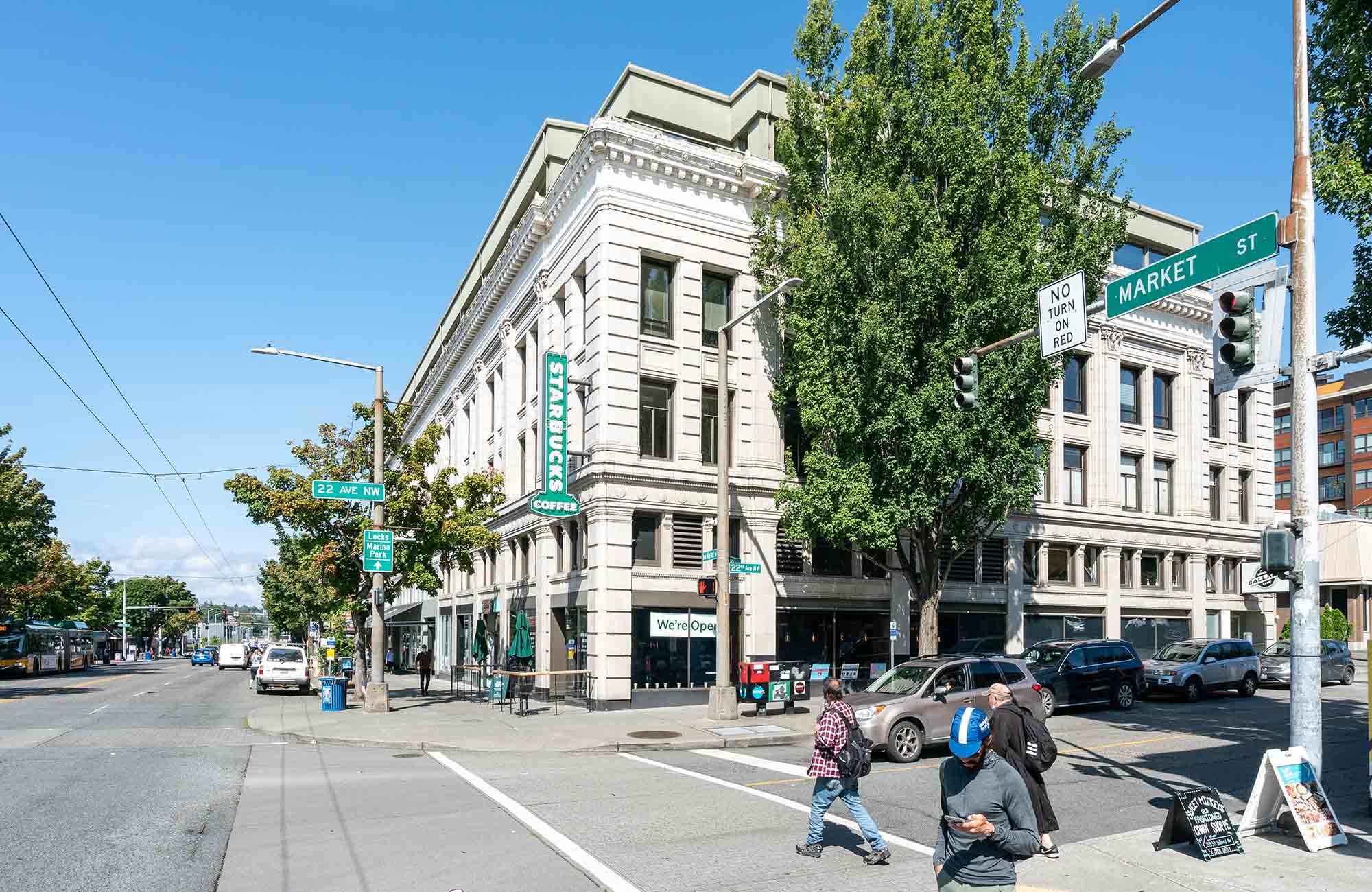 Historic corner building at NW Market Street and 22nd Ave NW in Ballard, Seattle, featuring a Starbucks and pedestrians crossing the intersection under clear blue skies.