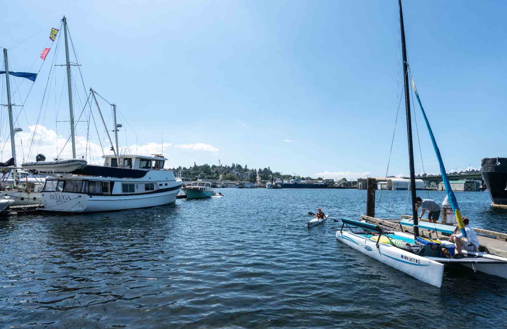 Boats and kayakers along the Ballard waterfront on a sunny day, with calm blue water, docked sailboats, and industrial buildings in the distance.