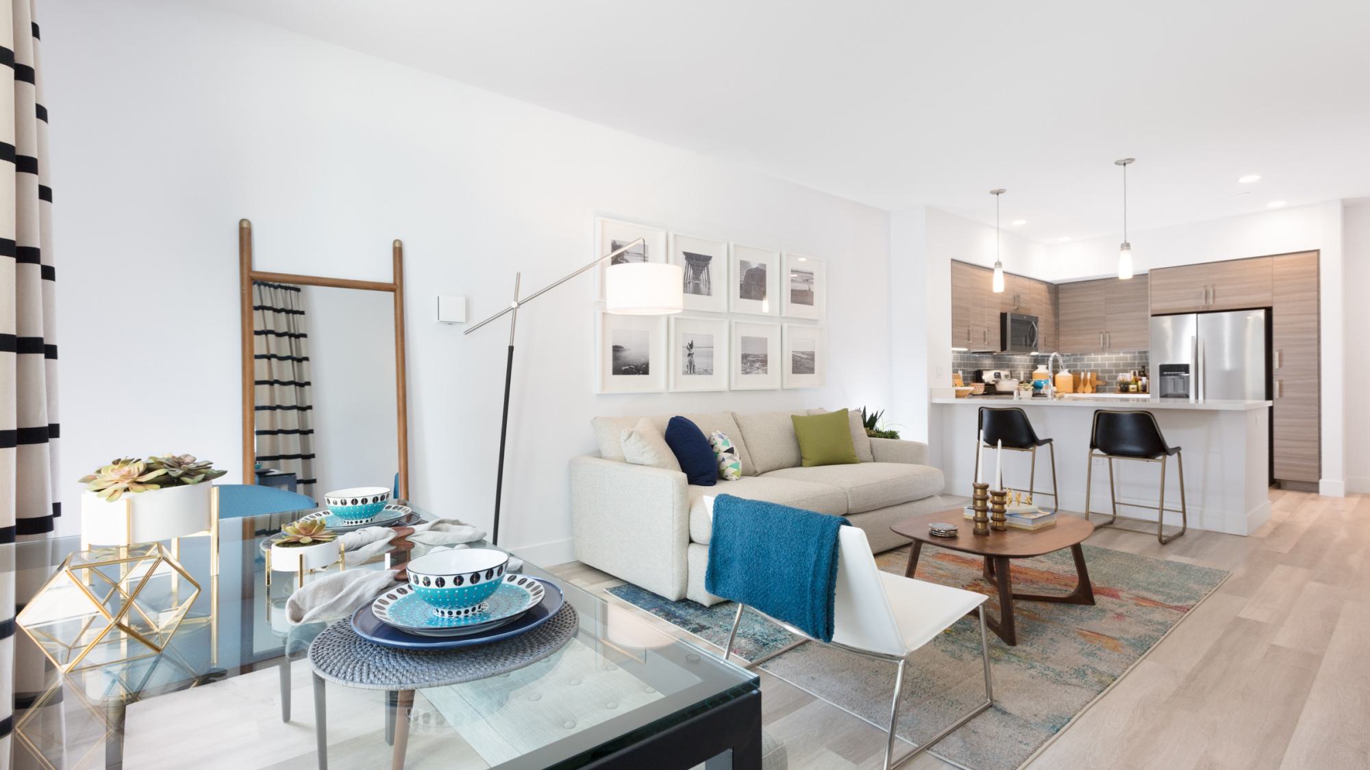 Modern open-concept apartment with a bright living room, glass dining table, and a sleek kitchen featuring wood cabinetry and stainless steel appliances.