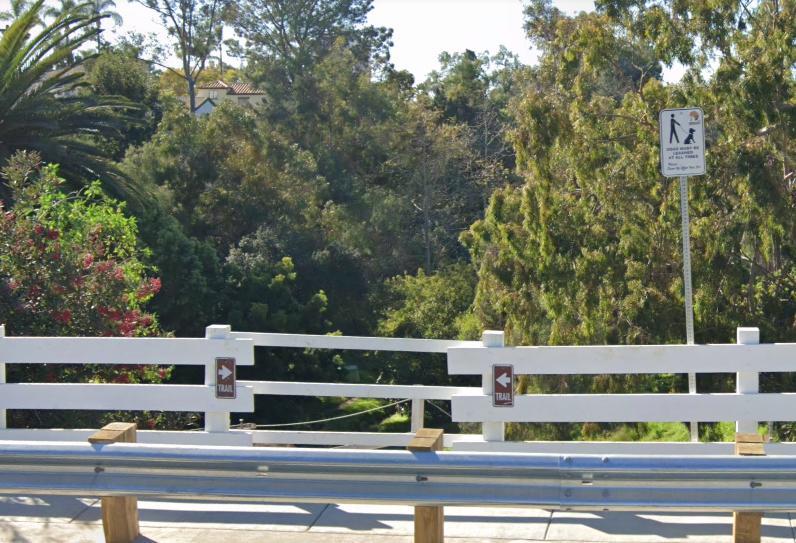 Entrance to Cabrillo Canyon Trail, marked by a white wooden fence with brown trail signs and a pedestrian crossing sign, leading into dense greenery and tall trees.