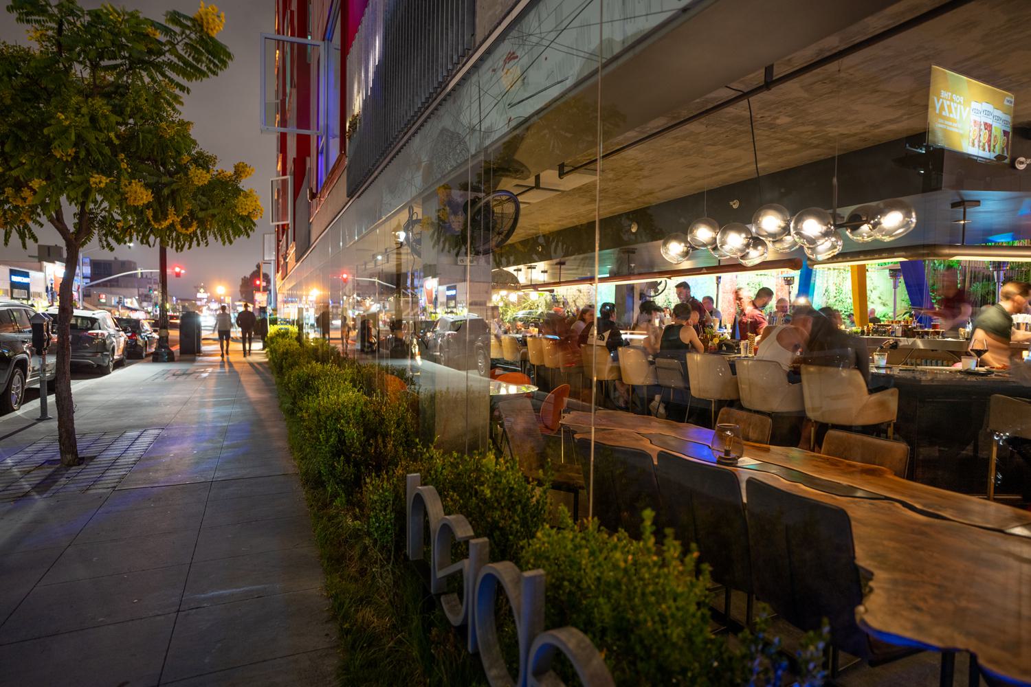 A lively Hillcrest street at night with a modern restaurant and bar visible through floor-to-ceiling glass walls, warm pendant lights illuminating the interior, and pedestrians walking along the tree-lined sidewalk outside.