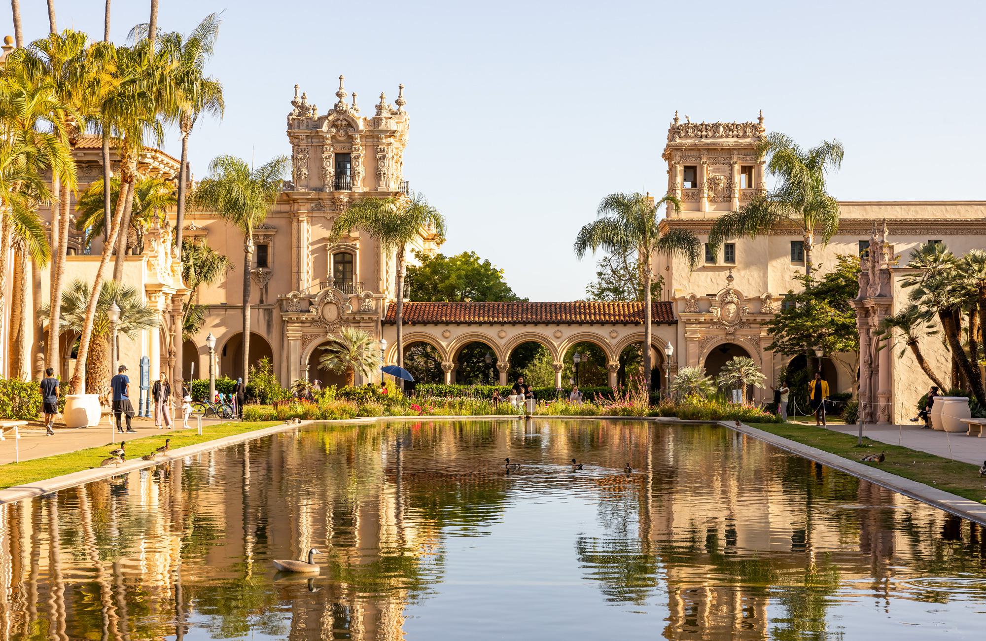The reflecting pool at Balboa Park in Hillcrest San Diego, surrounded by lush landscaping, palm trees, and ornate Spanish-Renaissance architecture with detailed facades and towers, under a clear afternoon sky.