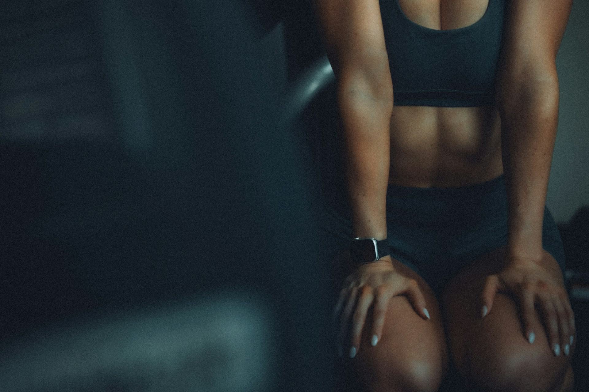 Close-up of a woman in a black sports bra and shorts sitting after a workout, showing toned abs and arms, with an Apple Watch on her wrist and soft, moody lighting.