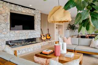 A modern club room featuring a stone fireplace with a mounted flat-screen TV, two acoustic guitars resting nearby, and a seating area with light gray sofas, warm wood accents, geometric accent pillows, and a large woven pendant light overhead. Greenery in planters softens the space, while art books and ceramic decor add a refined touch to the wooden coffee table.