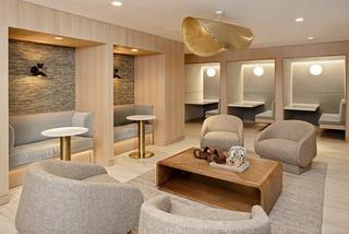 Modern coworking lounge at Denizen featuring soft neutral tones, curved lounge chairs around a wood coffee table, private work pods with built-in seating, and a sculptural gold pendant light overhead.