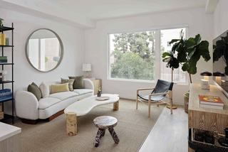 Bright apartment living room at Denizen with oversized windows, a curved white sofa, sculptural coffee table, modern accent chair, and a large fiddle-leaf fig plant adding greenery to the space.