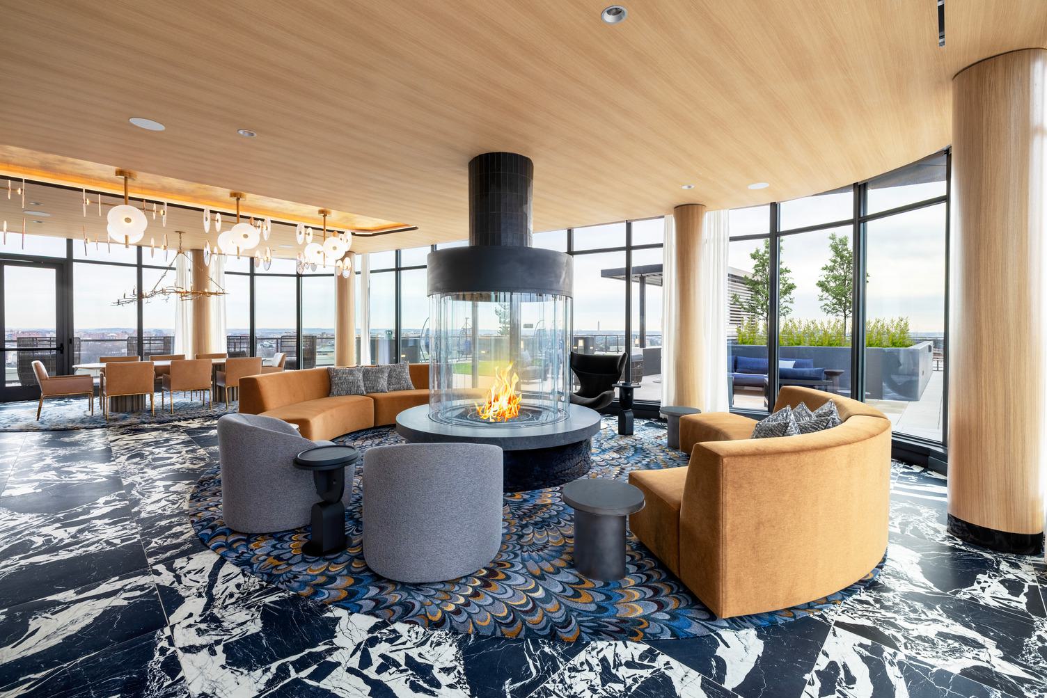The Observatory lounge at Zephyr in Woodley Park, DC, featuring a circular glass fireplace, plush seating, and floor-to-ceiling windows opening to panoramic views of the city skyline, including the Washington Monument at sunset.
