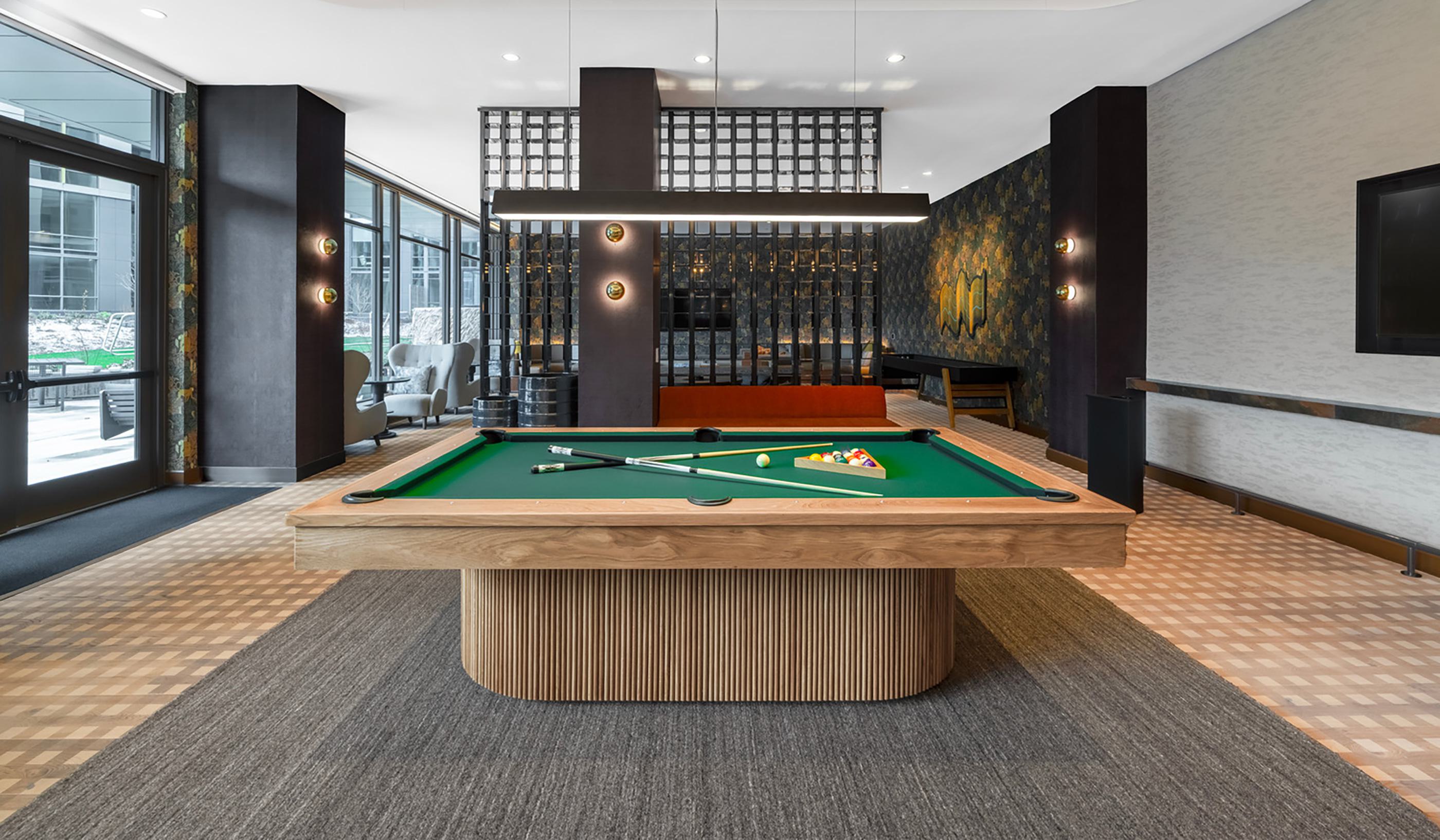 Game lounge at Zephyr in Woodley Park, DC, featuring a pool table, foosball table, bar-style seating, and cozy booths, with warm lighting and floor-to-ceiling windows overlooking landscaped outdoor areas.