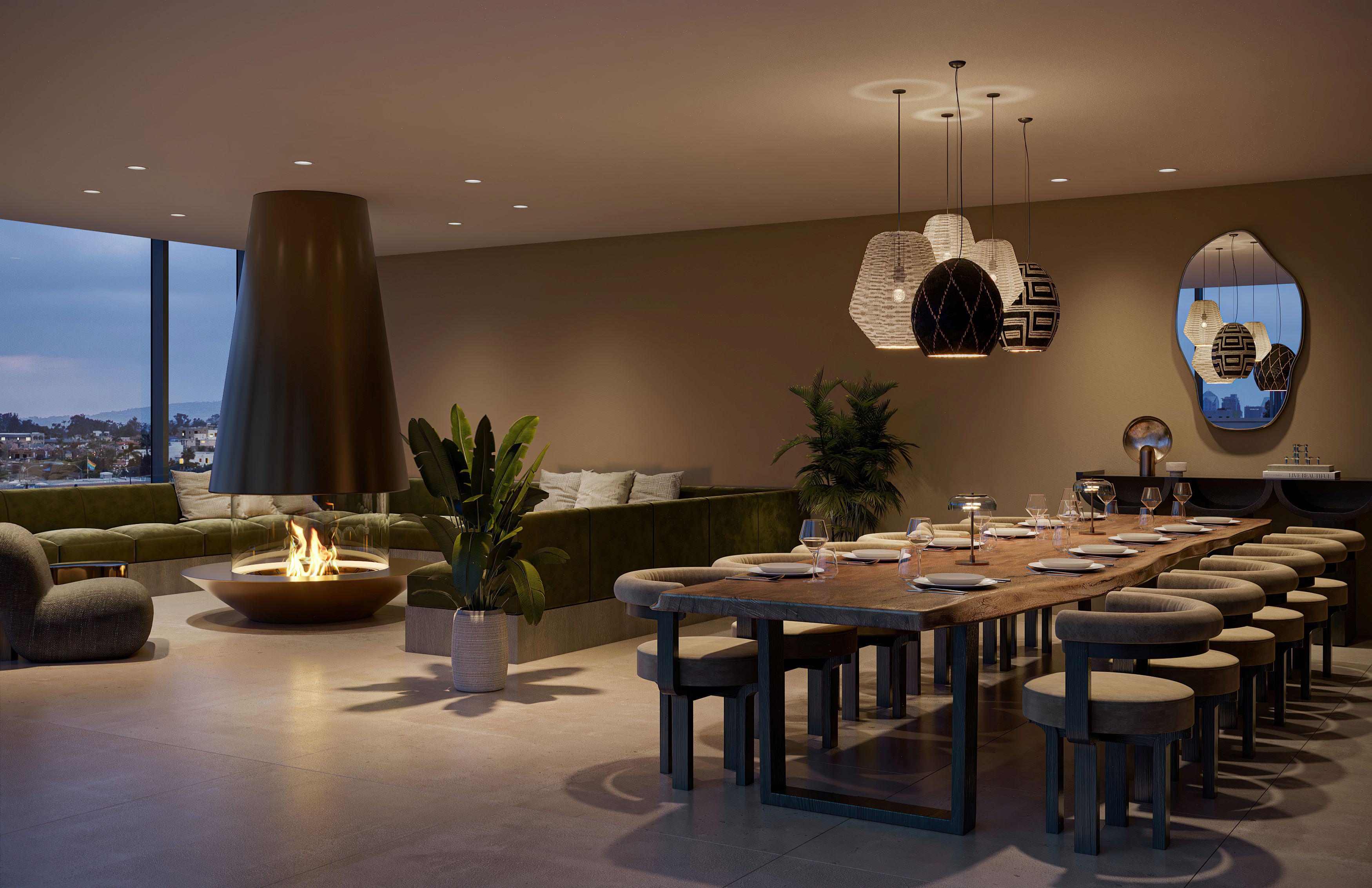 Rooftop lounge at Rowyn in Hillcrest, San Diego, with a modern suspended fireplace, deep green velvet sectional seating, and a long rustic wood dining table set for twelve. Overhead, sculptural pendant lights in black, white, and geometric patterns add a bold, artistic touch, while floor-to-ceiling windows frame twilight city views.