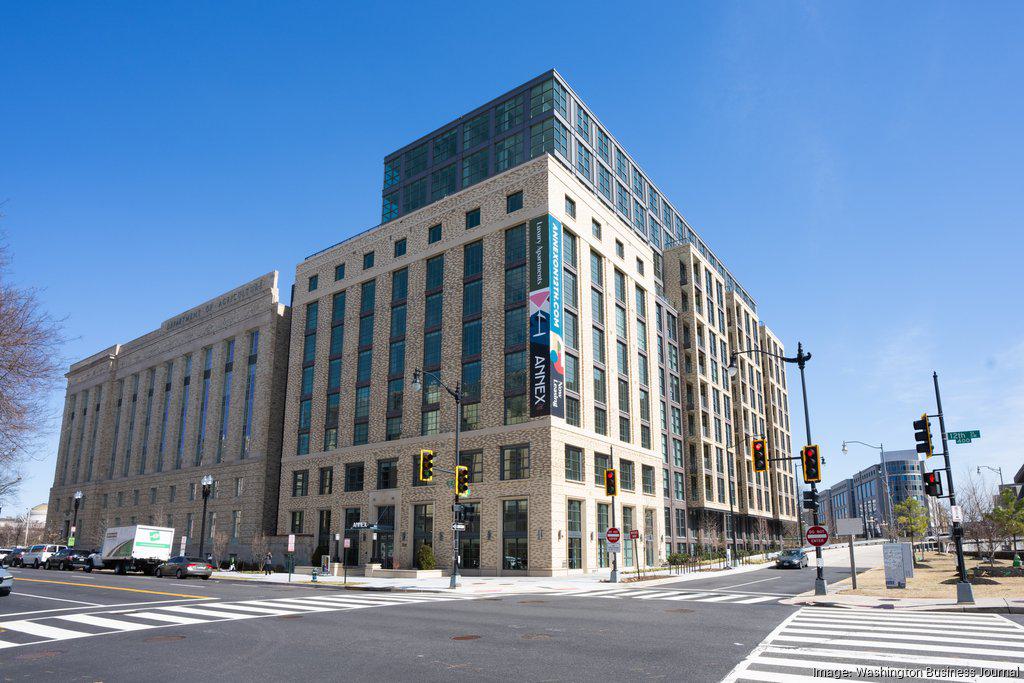 Annex on 12th, an adaptive reuse project in Southwest DC, featuring a historic brick façade integrated with a modern glass addition. The building stands on a corner with wide crosswalks, traffic lights, and clear blue skies, highlighting its transformation from a federal building into a contemporary mixed-use space.