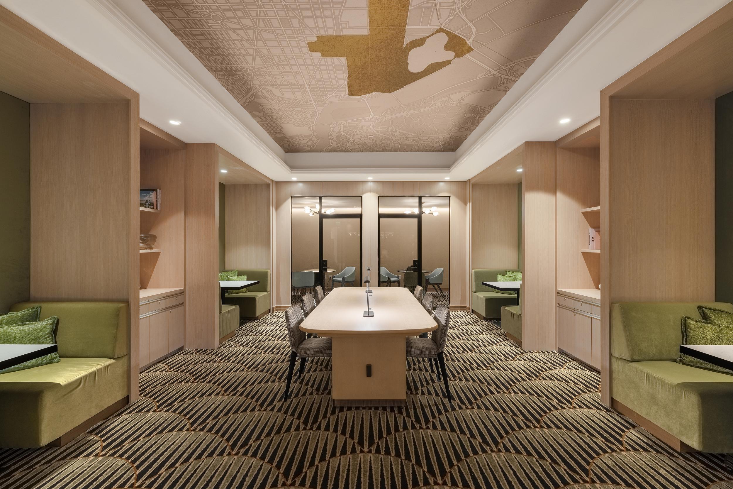 A refined coworking space at Annex on 12th with a central light-wood table surrounded by chairs, semi-private green-upholstered booths along both sides, and a ceiling featuring a gold-accented map design. Patterned carpet adds texture, and glass doors at the far end lead into an adjoining room with additional seating.
