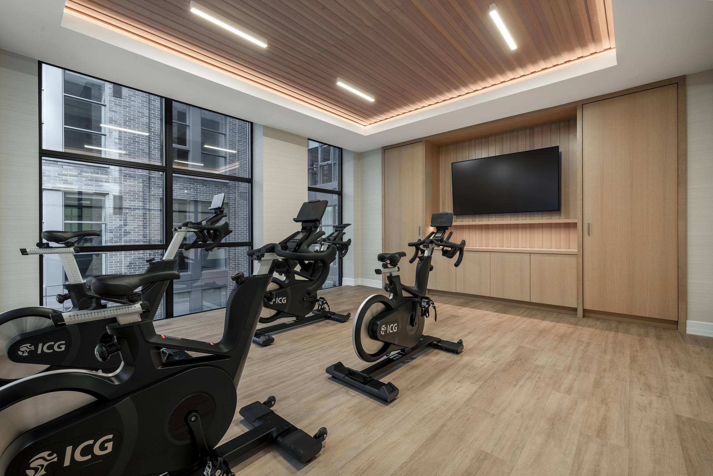 A sleek spin studio at Annex on 12th with three ICG stationary bikes, a wall-mounted TV, wood-paneled storage, and a slatted wood ceiling with recessed lighting. Large windows bring in natural light and views of the building’s brick exterior.