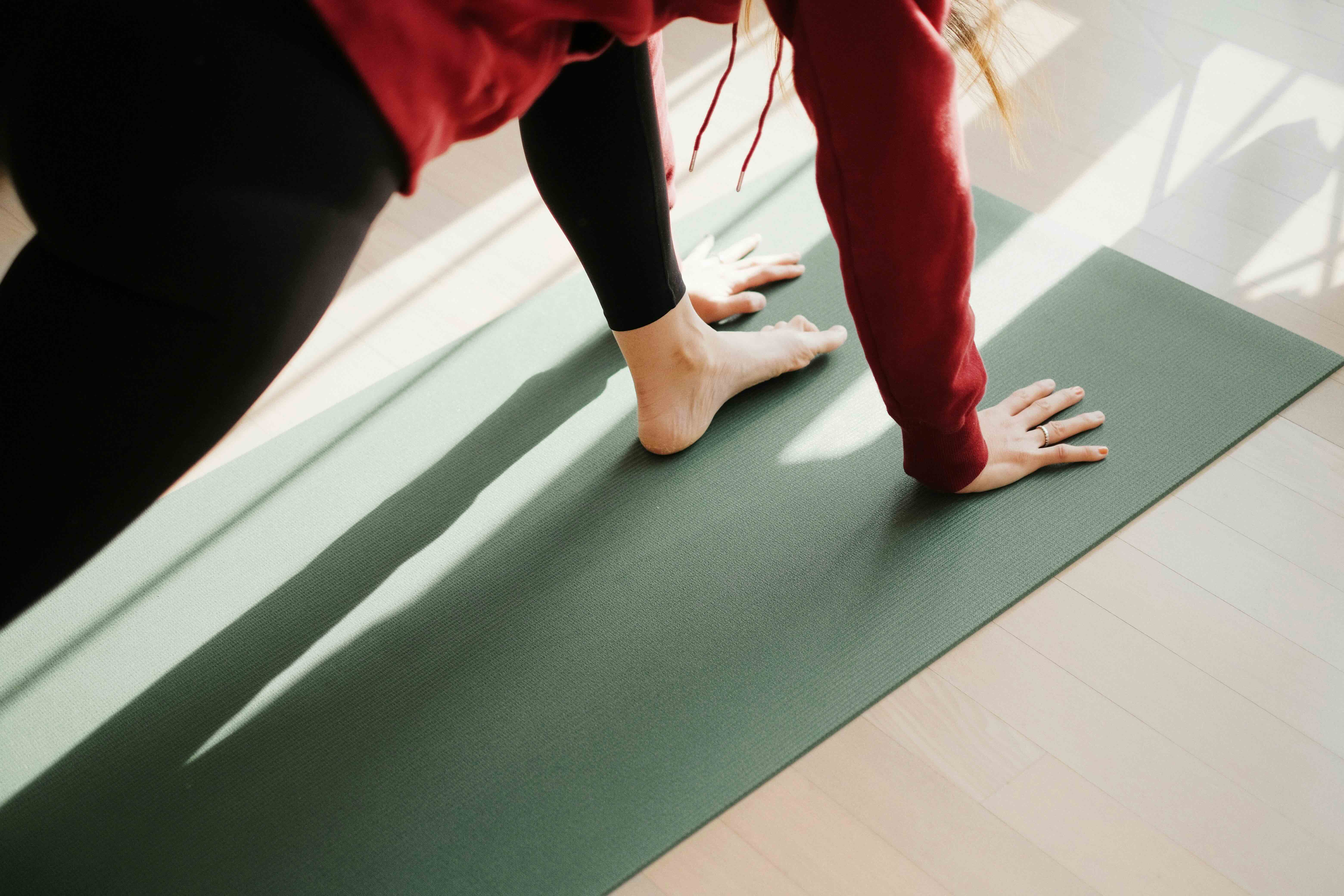 A close-up of a person in a yoga pose on a sage green mat, wearing a red sweatshirt and black leggings, with soft natural light and shadows cast across light wood flooring.
