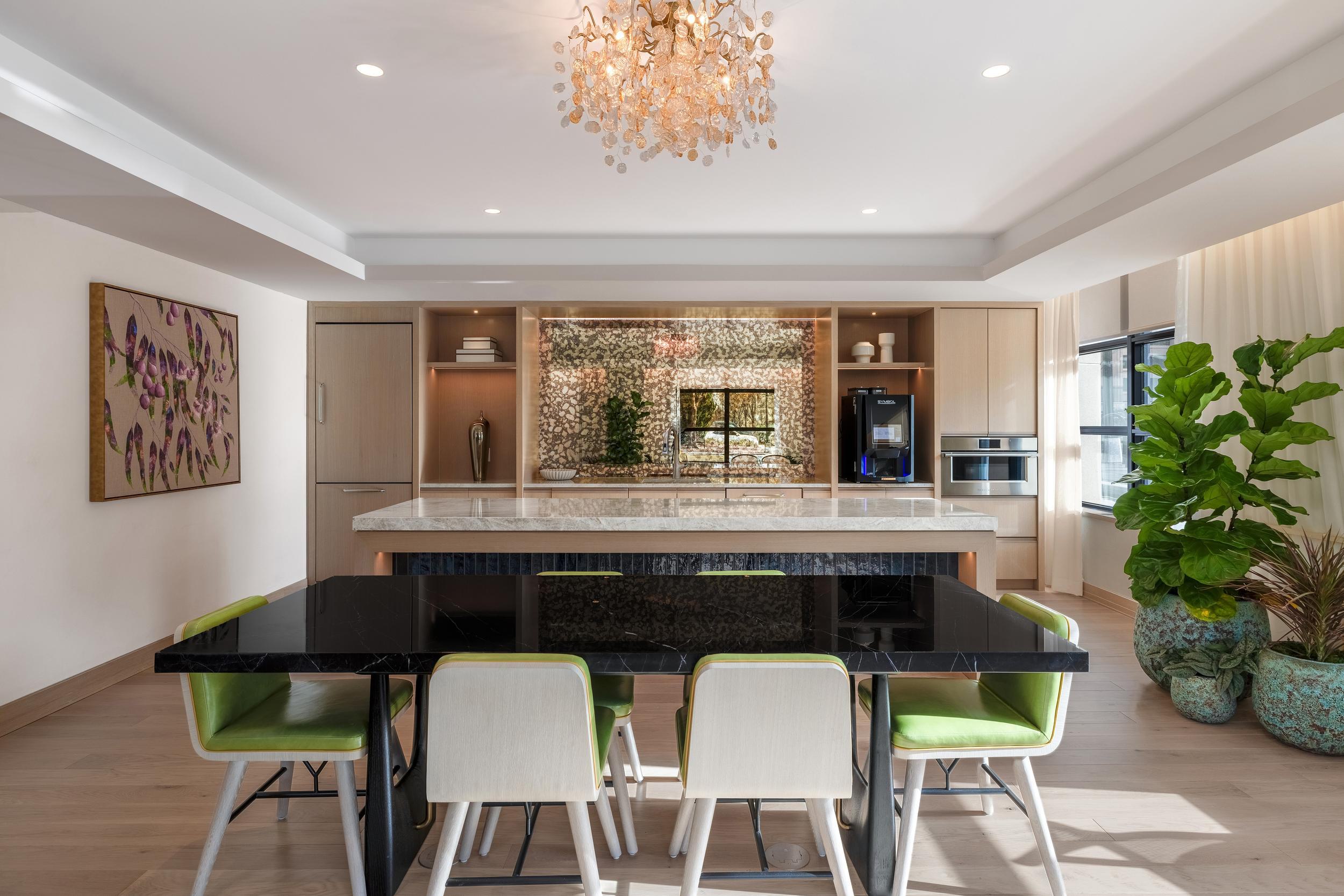 Elegant orangery space featuring a black dining table with green-accented chairs, a marble-topped island, light wood cabinetry, and a mirrored backsplash with warm lighting under a modern chandelier.
