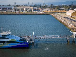 Passengers boarding a San Francisco Bay Ferry at the Seaplane Lagoon Terminal in Alameda, with the pier stretching over the water and the former naval air station and industrial buildings visible in the background.