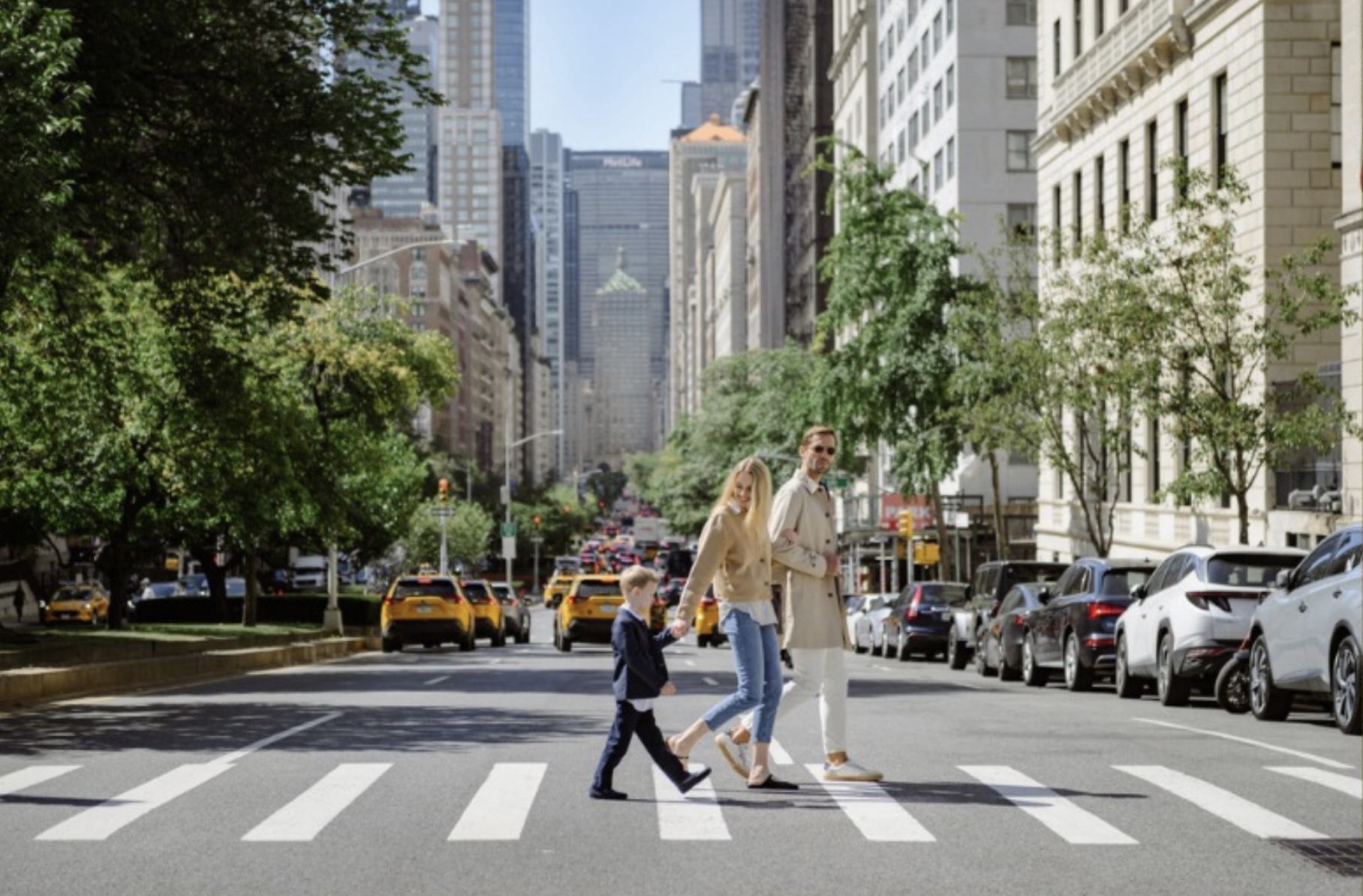 A stylish family crosses a tree-lined Upper East Side street in New York City, with yellow taxis and Midtown skyscrapers rising in the background.