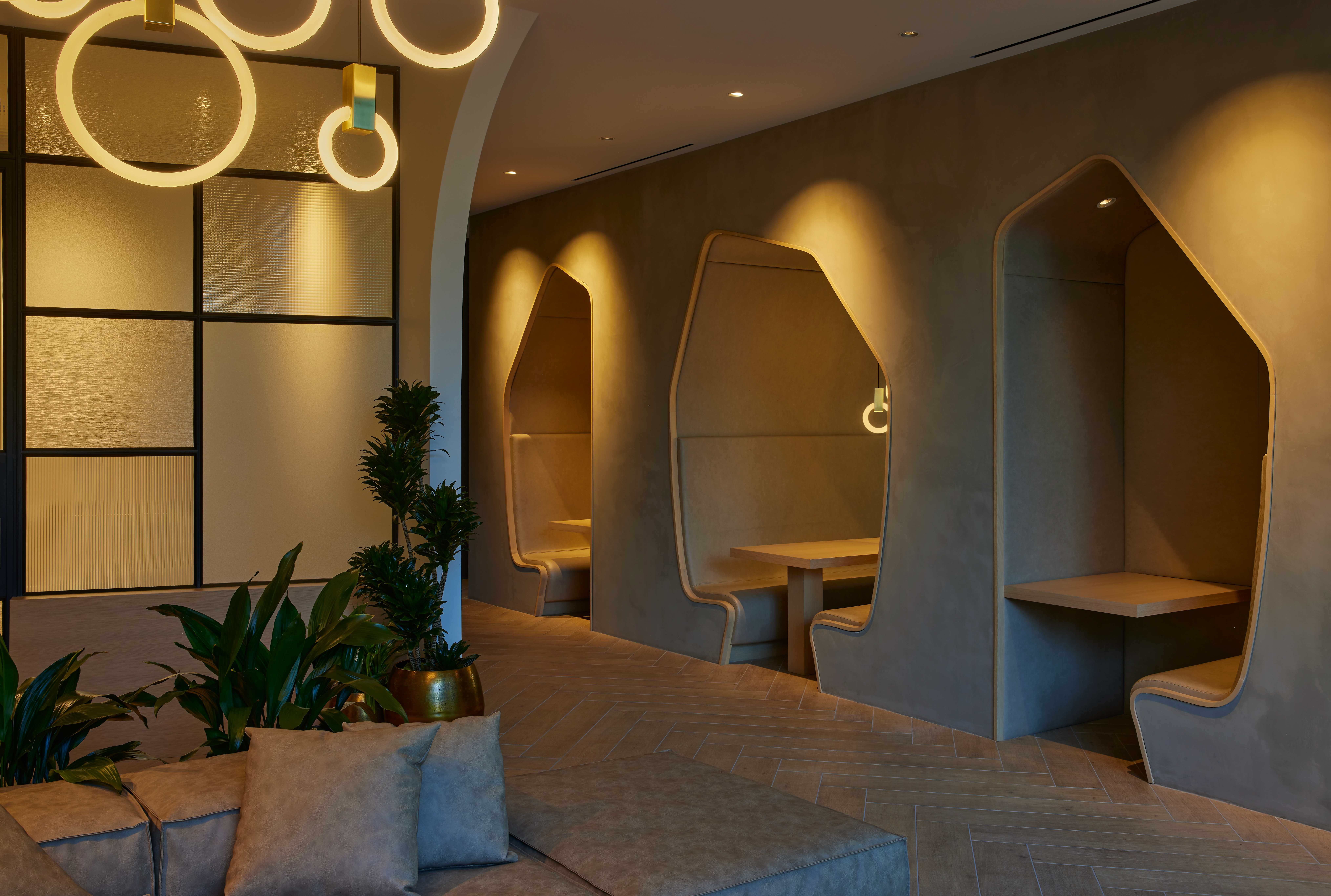 Modern coworking space with curved booth seating, integrated lighting, circular pendant fixtures, textured glass panels, and potted plants