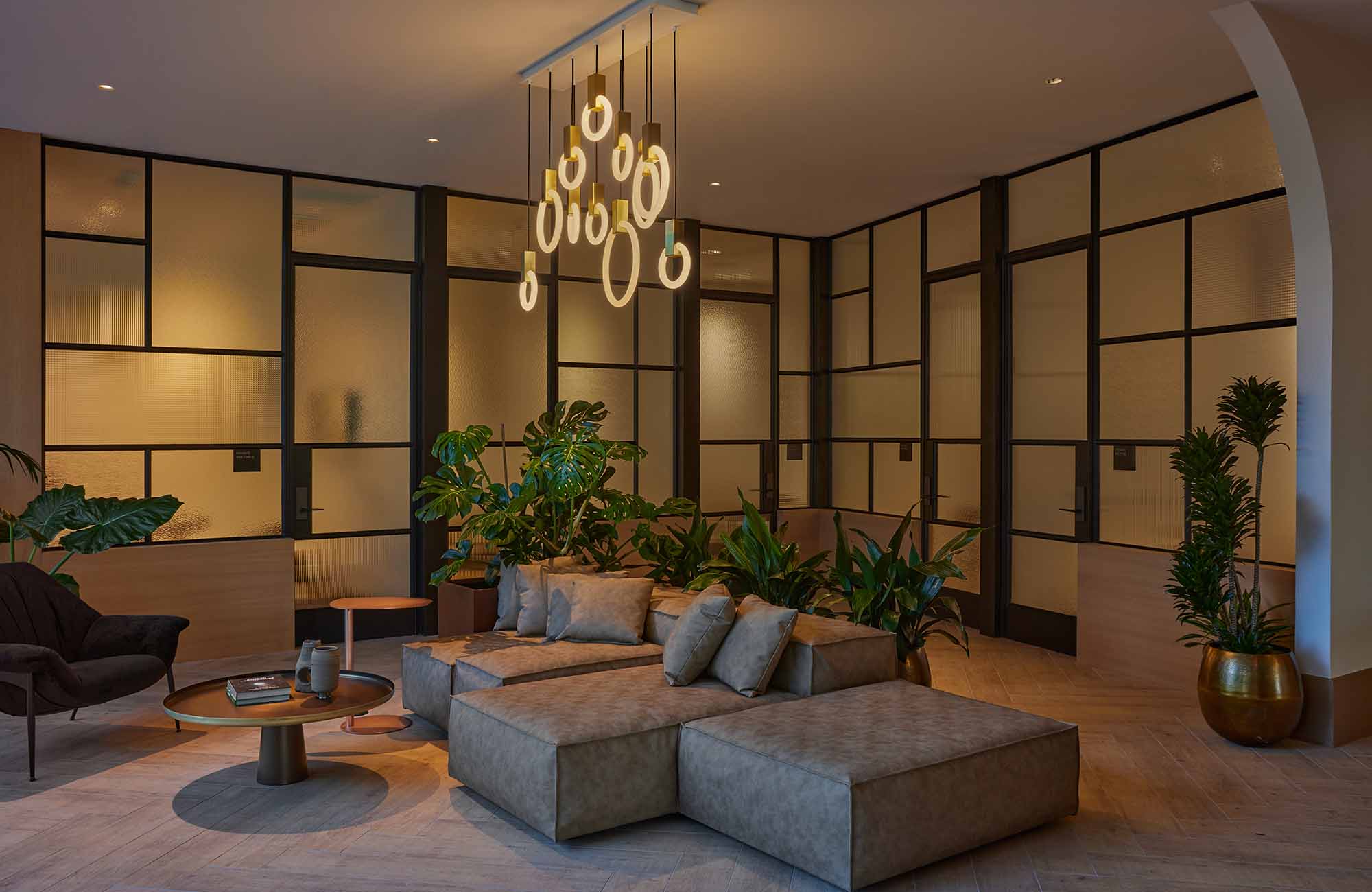 Contemporary lounge area with gray sectional seating, circular pendant lighting, floor-to-ceiling textured glass panels, and abundant greenery