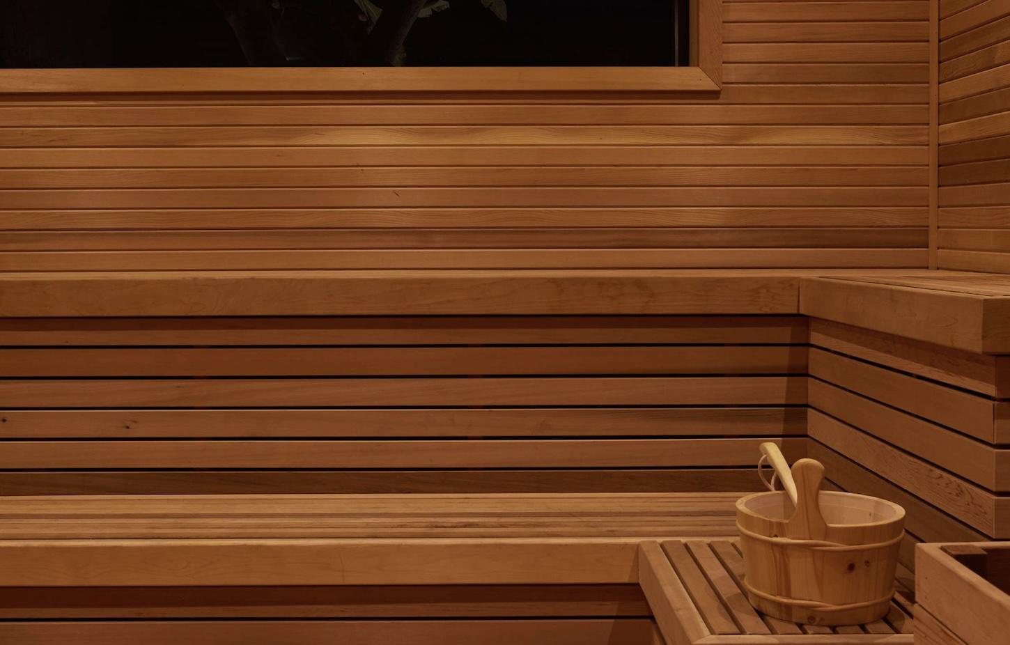Interior of wooden sauna with horizontal wood paneling, tiered bench seating, window with greenery view, and traditional wooden bucket and ladle