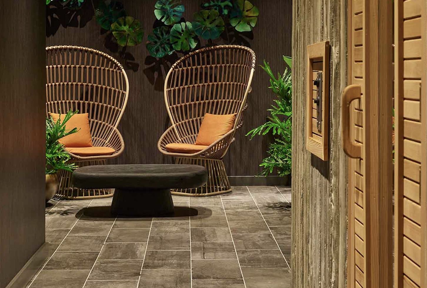 Sauna relaxation area with two woven rattan chairs with orange cushions, dark wood paneling, natural stone flooring, and potted plants