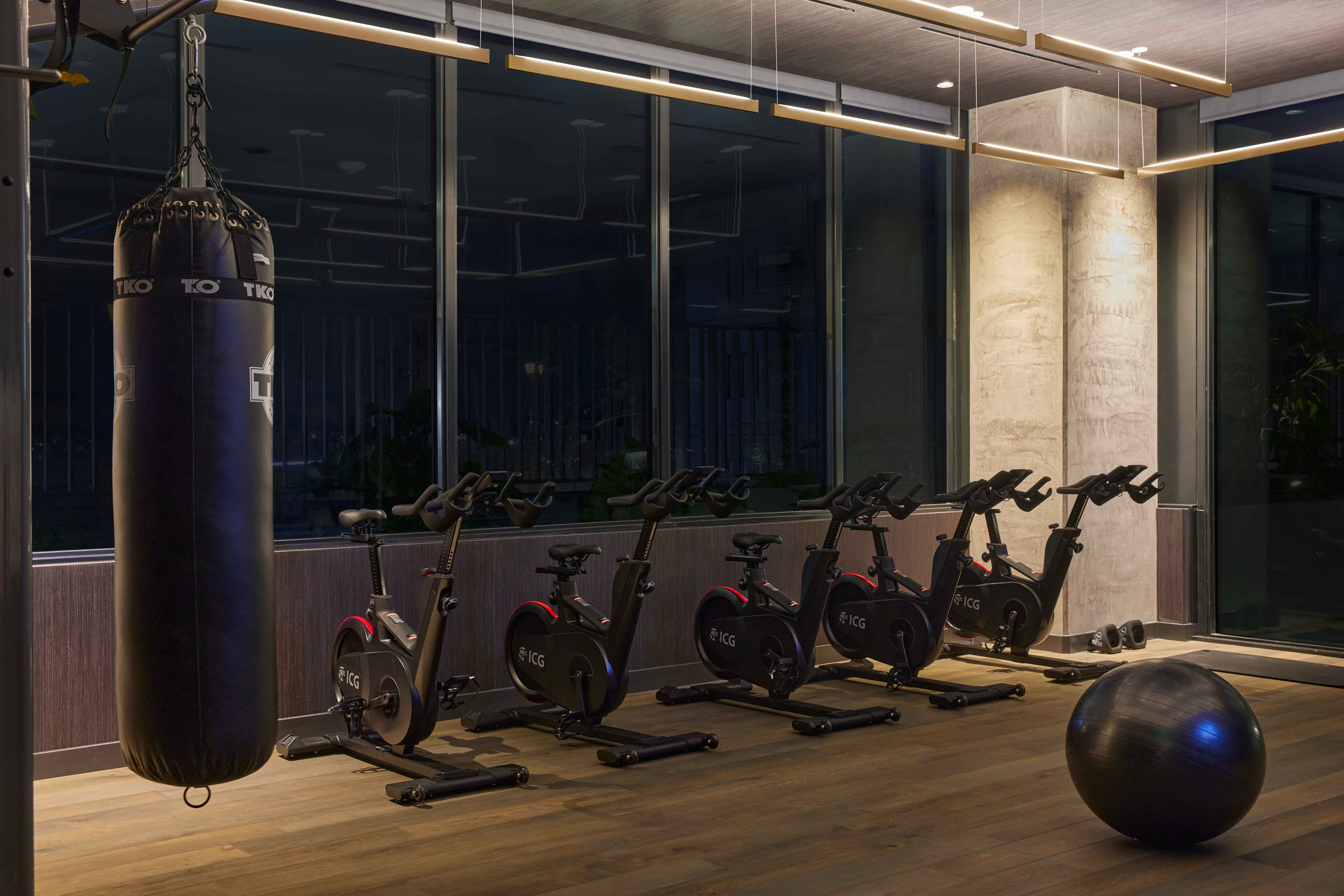 Spin studio with row of black Echelon bikes facing floor-to-ceiling windows, suspended TKO punching bag, and wood flooring