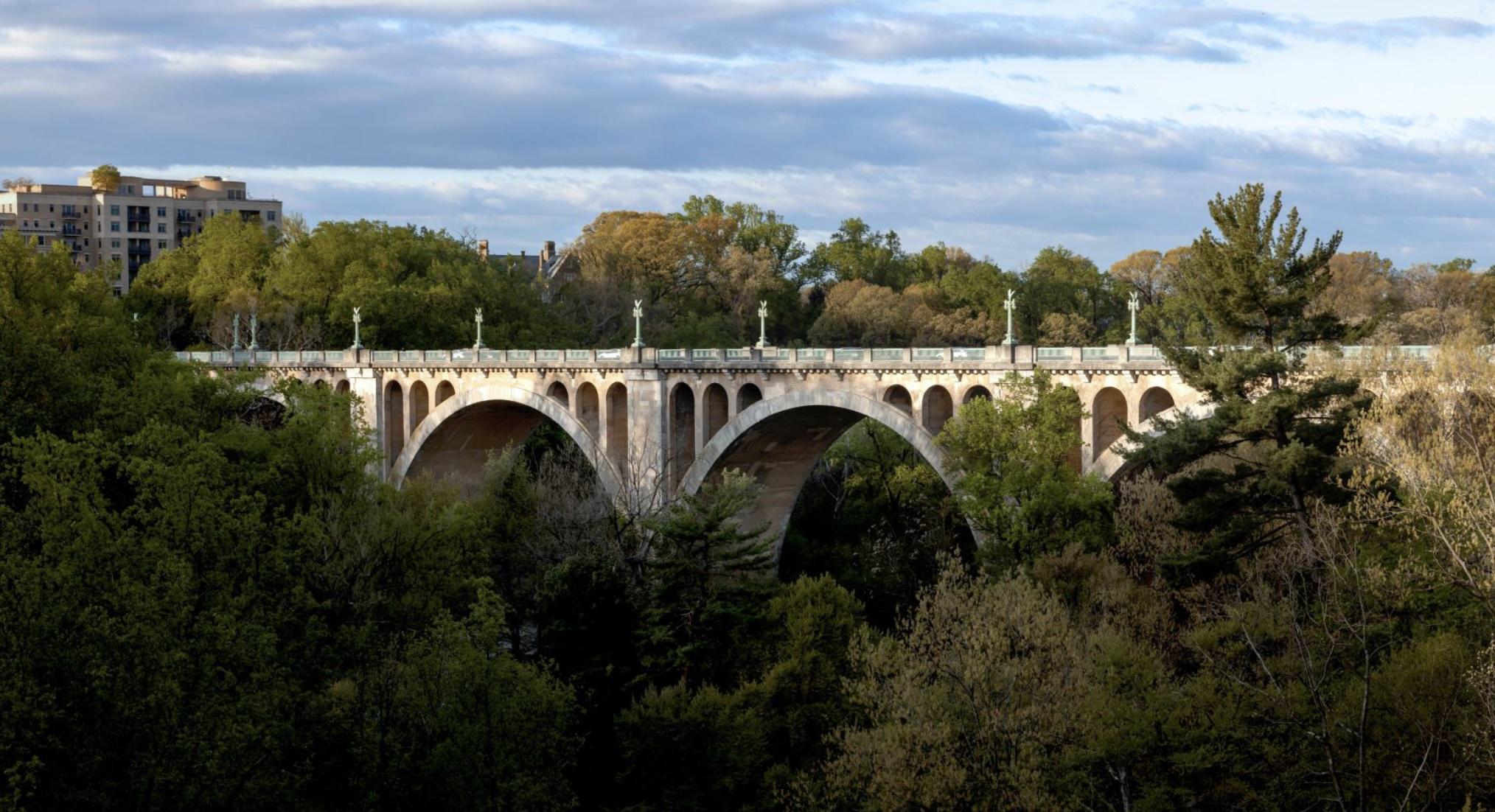 Connecticut Avenue Bridge that connects Woodley Park to Dupont Circle with multiple arches spans across a tree-filled valley in autumn. The bridge features classical architecture with decorative balustrades and lamp posts. Fall foliage in golden and green tones fills the gorge below, while modern apartment buildings are visible in the background under a cloudy sky.