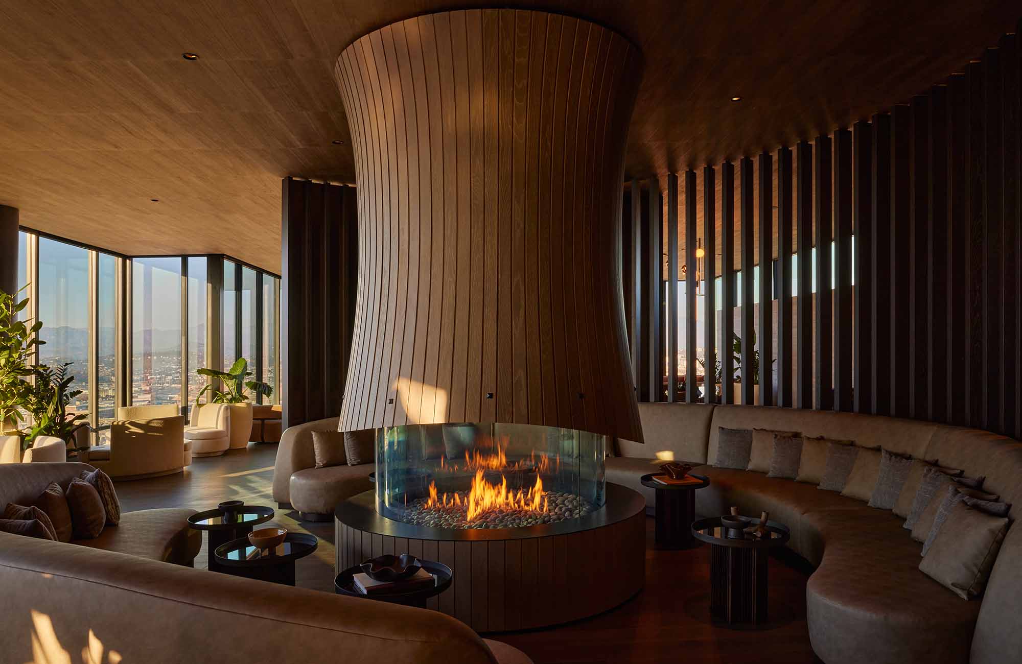 Sophisticated rooftop lounge with sculptural fireplace, curved banquette seating, textured golden bar, and floor-to-ceiling windows offering LA views. Features warm wood and stone finishes with vertical privacy screens.