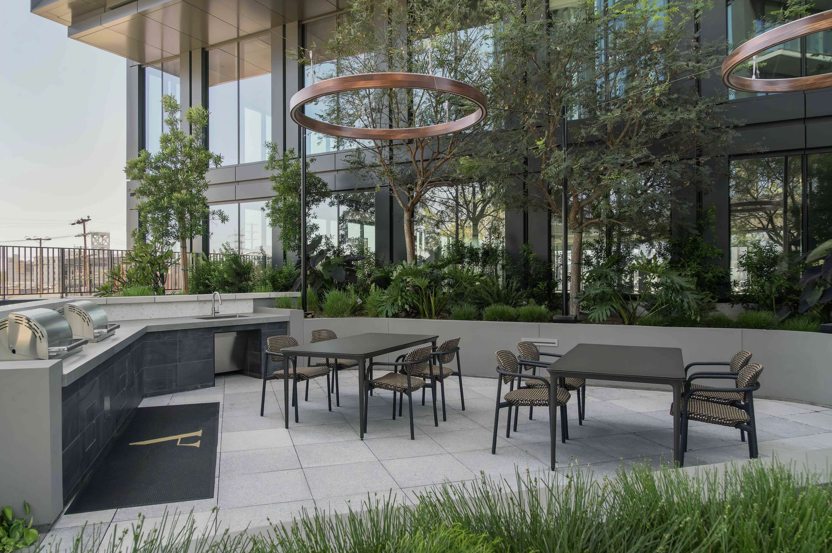 Modern rooftop terrace featuring an outdoor kitchen with concrete countertops and dark cabinetry, two dining tables with woven chairs, and distinctive copper circular light fixtures suspended above lush planted areas.