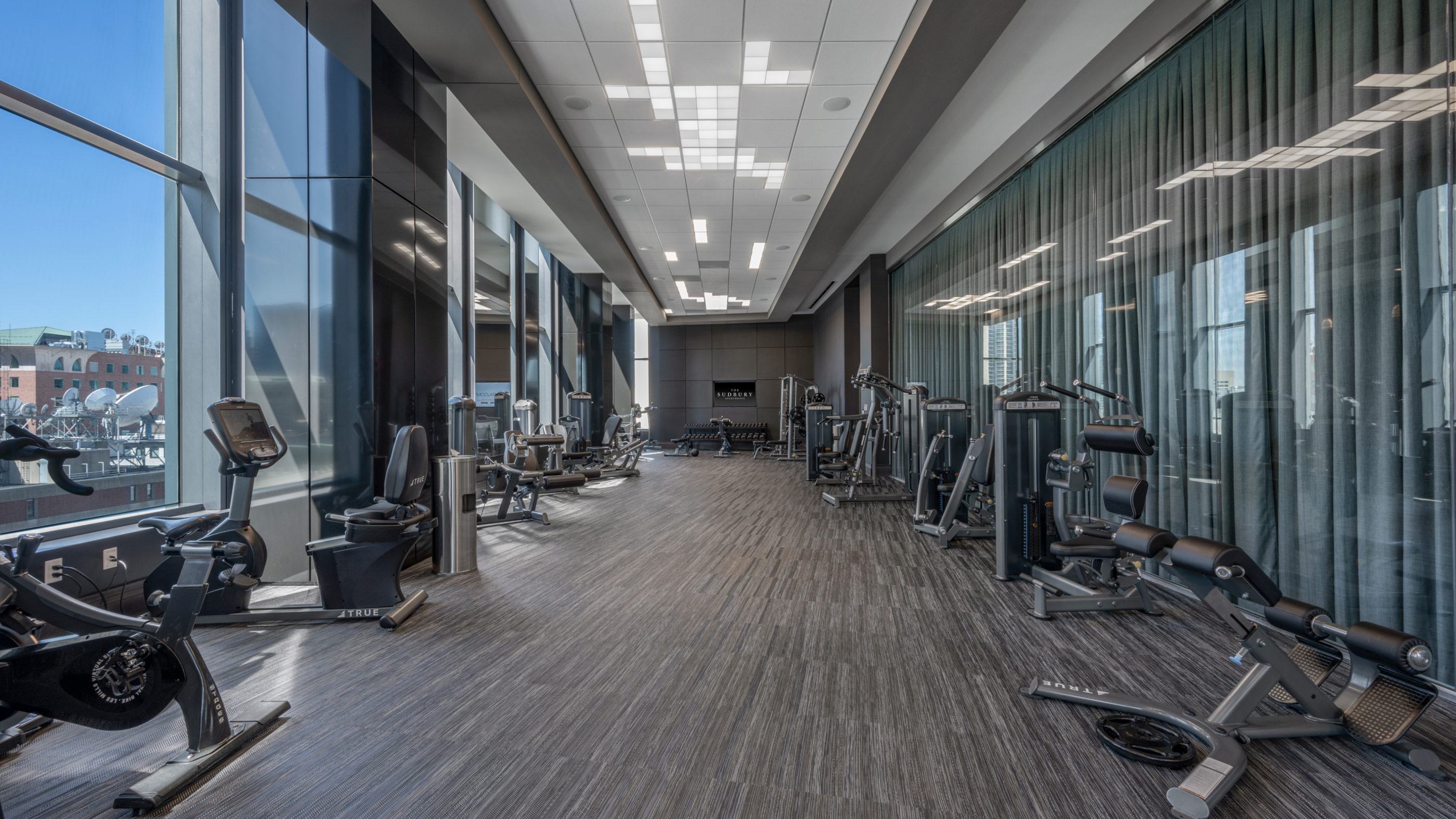 Expansive fitness center with floor-to-ceiling windows overlooking Boston, featuring cardio machines, weight benches, and strength-training equipment in a bright, open layout.