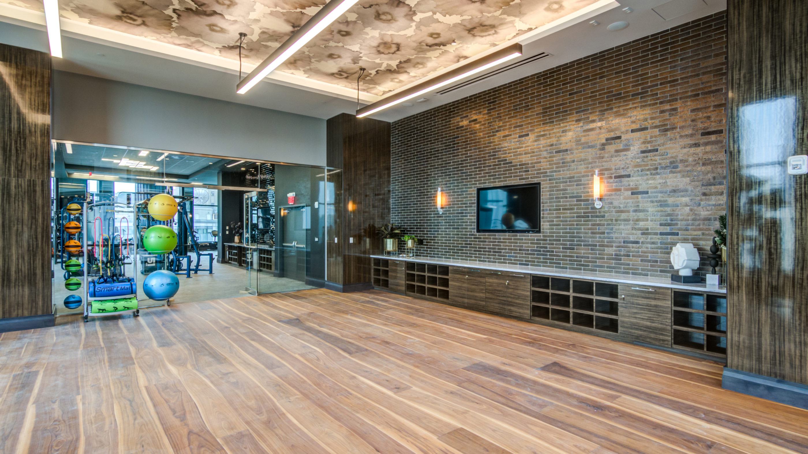 Spacious yoga and Pilates studio with warm wood floors, a brick accent wall, built-in cabinetry, and racks holding colorful stability balls and resistance equipment.