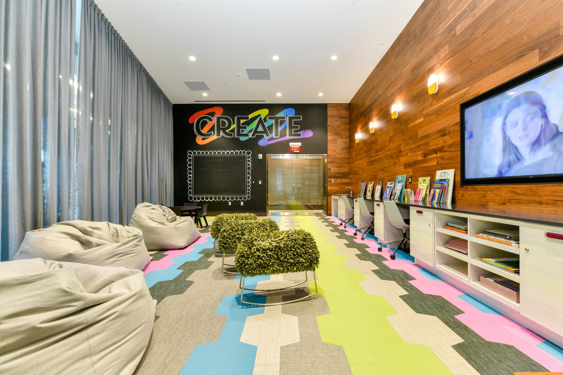 Colorful kids’ playroom with patterned flooring, wood-paneled wall, desks with books and art supplies, and a large screen for entertainment.
