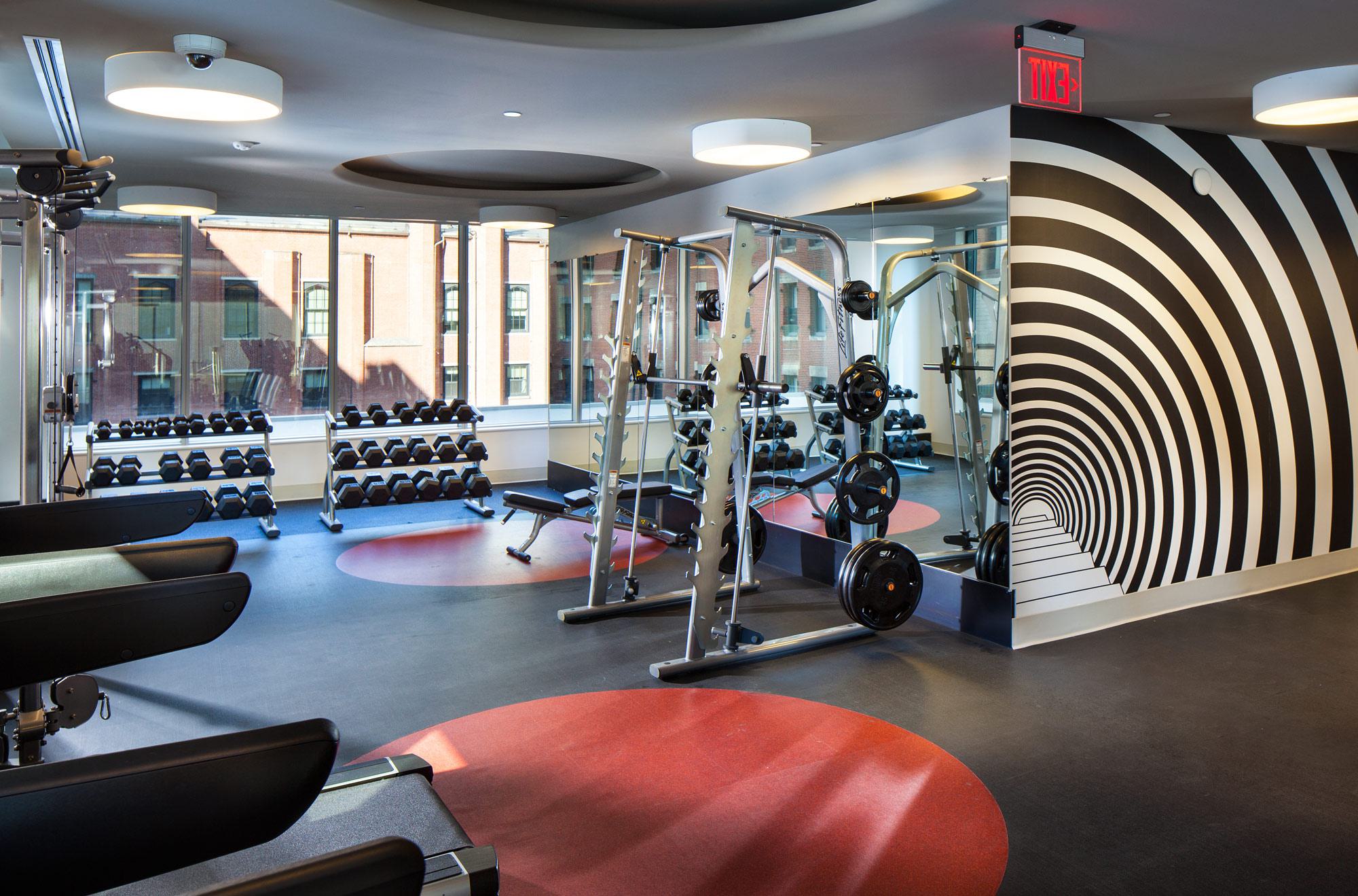 Strength training area with free weights, benches, and a squat rack, set against mirrored walls and bold black-and-white graphic wall art.