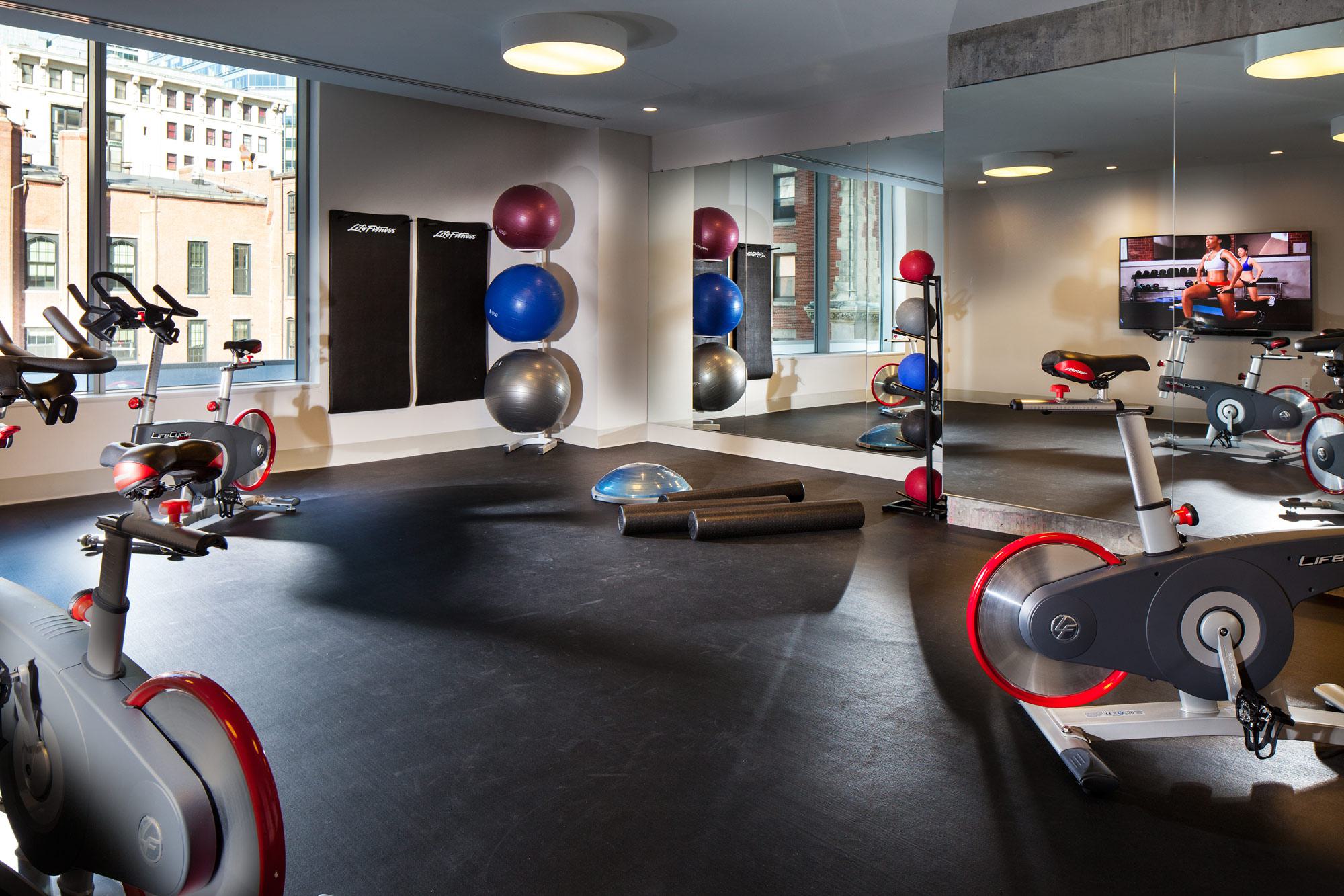 Spin studio with stationary bikes, mirrored walls, large windows, and neatly arranged equipment including medicine balls, stability balls, a BOSU trainer, and foam rollers.
