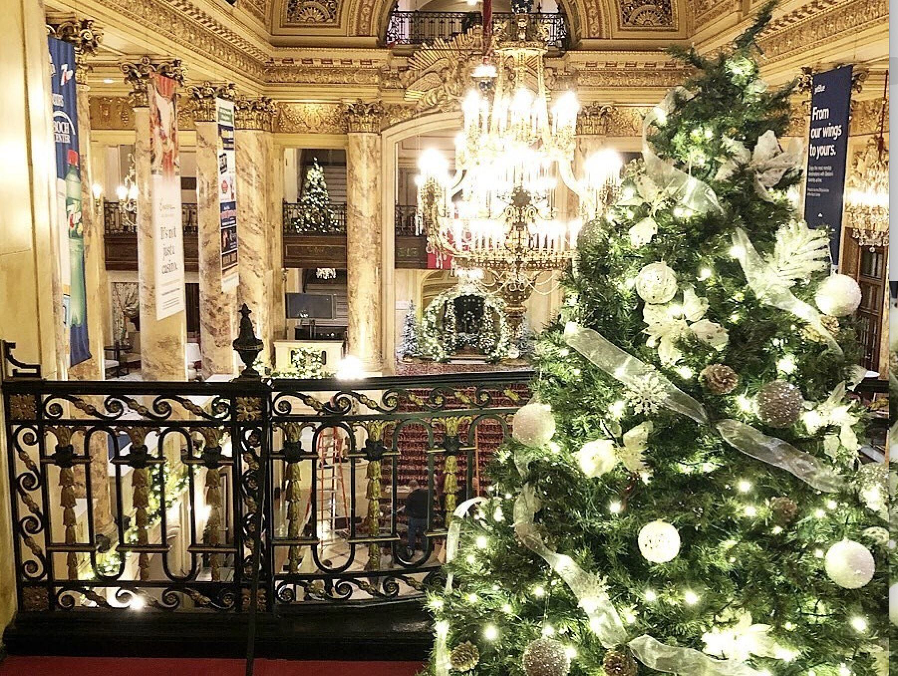 A grand holiday scene inside Boston’s Wang Theatre, with a sparkling Christmas tree decorated in white and gold ornaments in the foreground, glowing under warm chandelier light. Marble columns, ornate gilded details, and additional trees adorned with lights frame the elegant historic theater interior.