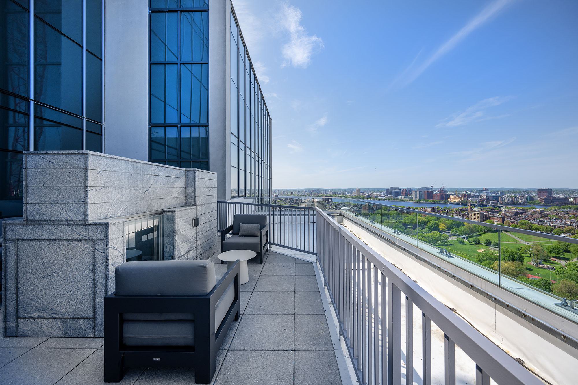 Elevated terrace at Luka on the Common with cushioned seating beside a stone fireplace, opening to panoramic views of Boston Common, the Charles River, and the city skyline beyond.
