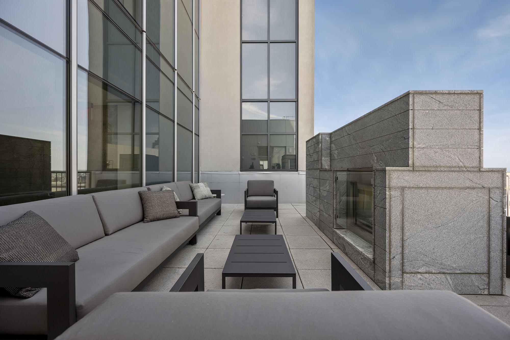 Outdoor terrace at Luka on the Common with long gray sofas, armchairs, and black tables arranged beside a sleek stone fireplace against the glass façade of the building.