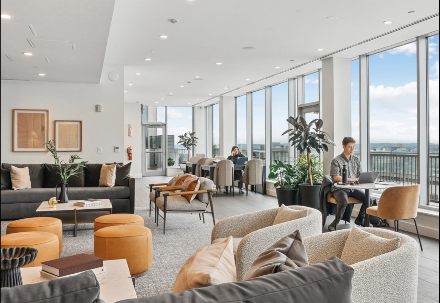 Light-filled sky lounge with cozy seating, leather ottomans, and café tables by tall windows where residents work while overlooking the city skyline.