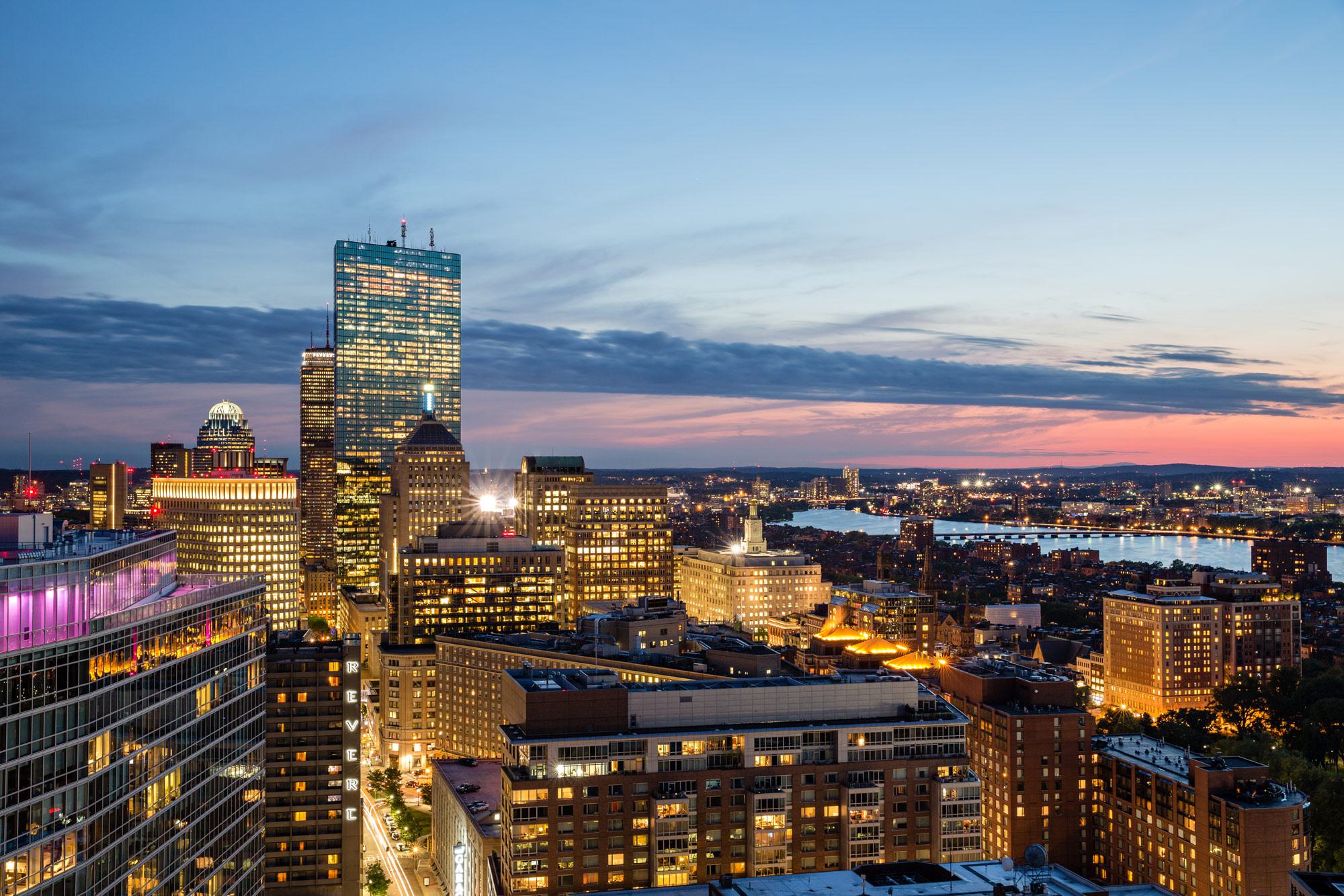Panoramic view of downtown Boston at dusk, with the Prudential Tower and surrounding high-rises lit against the evening sky, overlooking the Charles River.