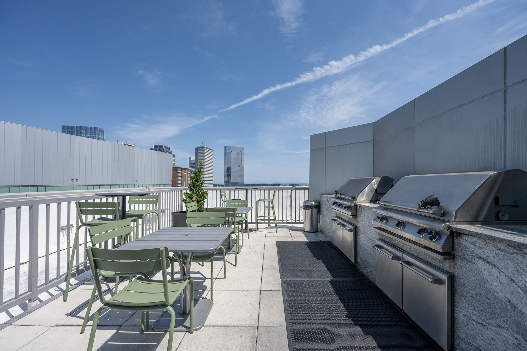 Outdoor grilling terrace at Luka on the Common with stainless steel gas grills, stone counters, and green café seating overlooking the Boston skyline.