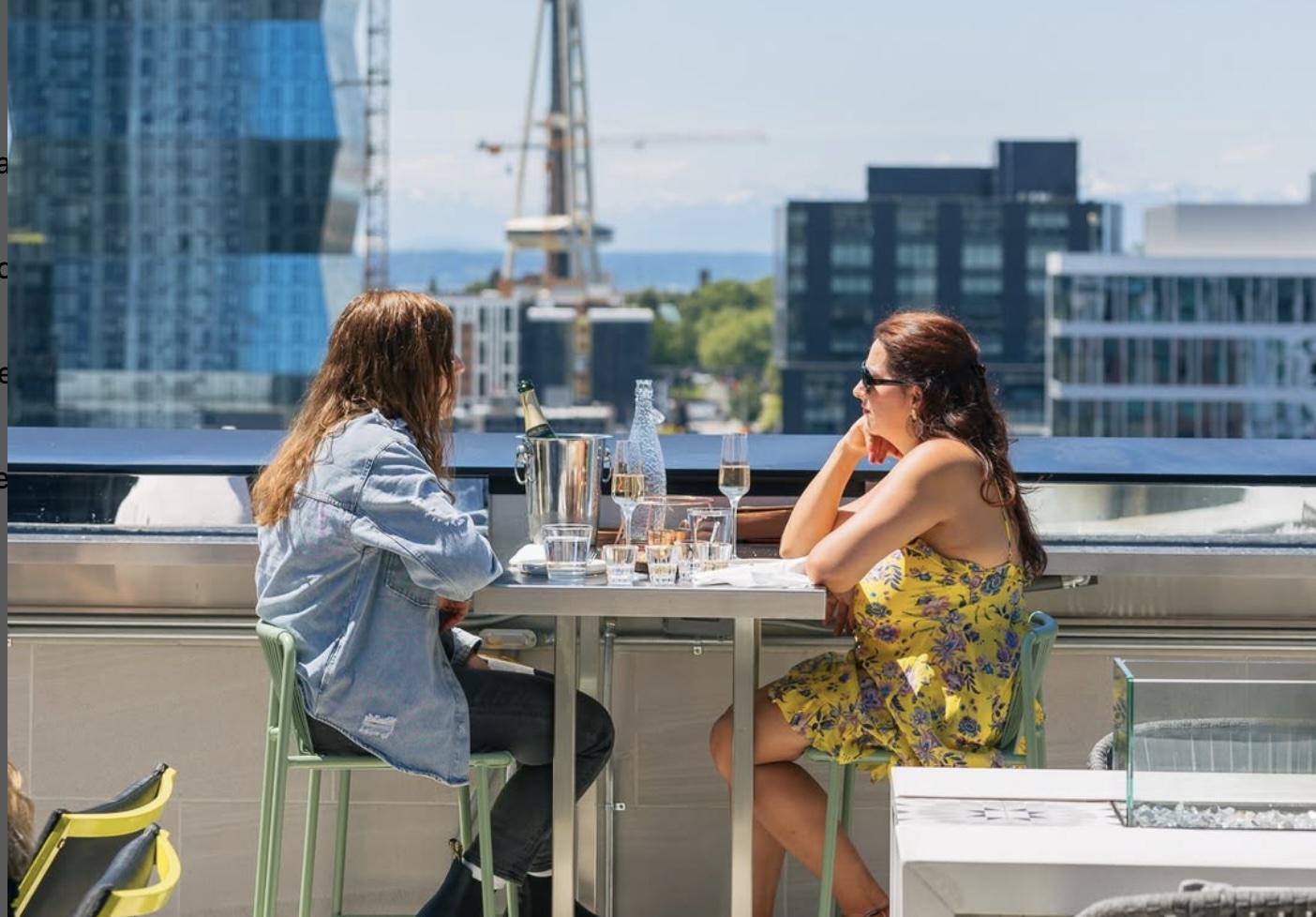 Two women sit at a high-top table on a rooftop terrace at Altitude, sharing champagne from a bottle in an ice bucket. One woman wears a denim jacket, the other a yellow floral dress and sunglasses. Modern Seattle buildings and construction cranes are visible in the background under a clear blue sky.