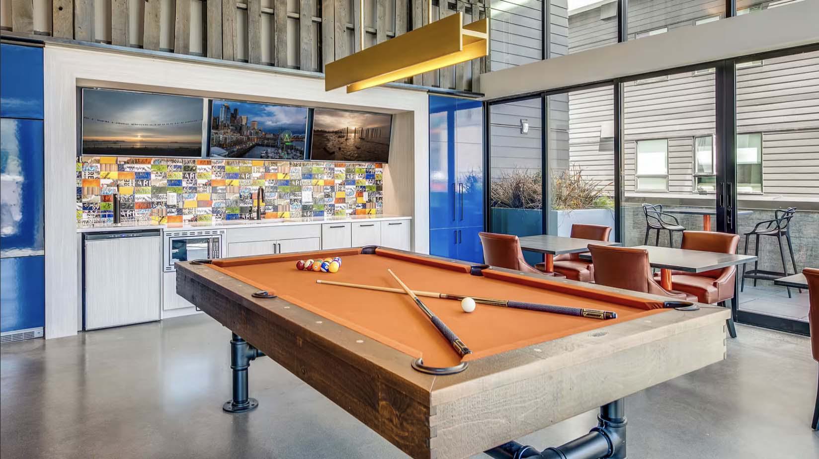 Modern lounge at Chroma with an orange-felt pool table in the center, leather chairs with café tables by floor-to-ceiling windows, and a colorful tiled backsplash with mounted city photos above a built-in bar area.