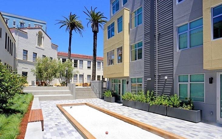 Outdoor bocce court at Alchemy framed by modern apartments, palm trees, planters, and seating areas for residents to gather.