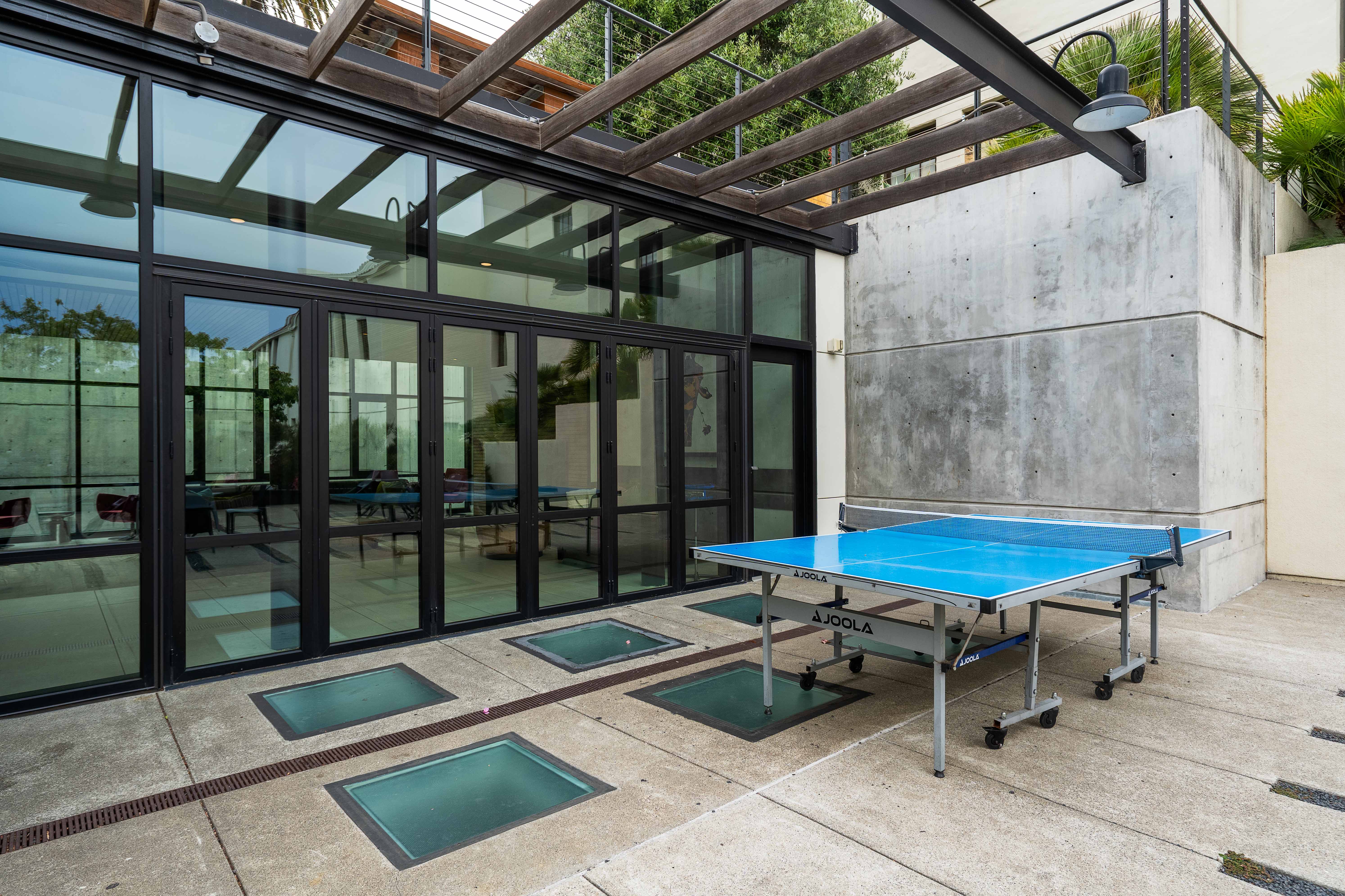 Outdoor ping pong table at Alchemy set on a concrete terrace beside floor-to-ceiling glass doors and a slatted pergola overhead.